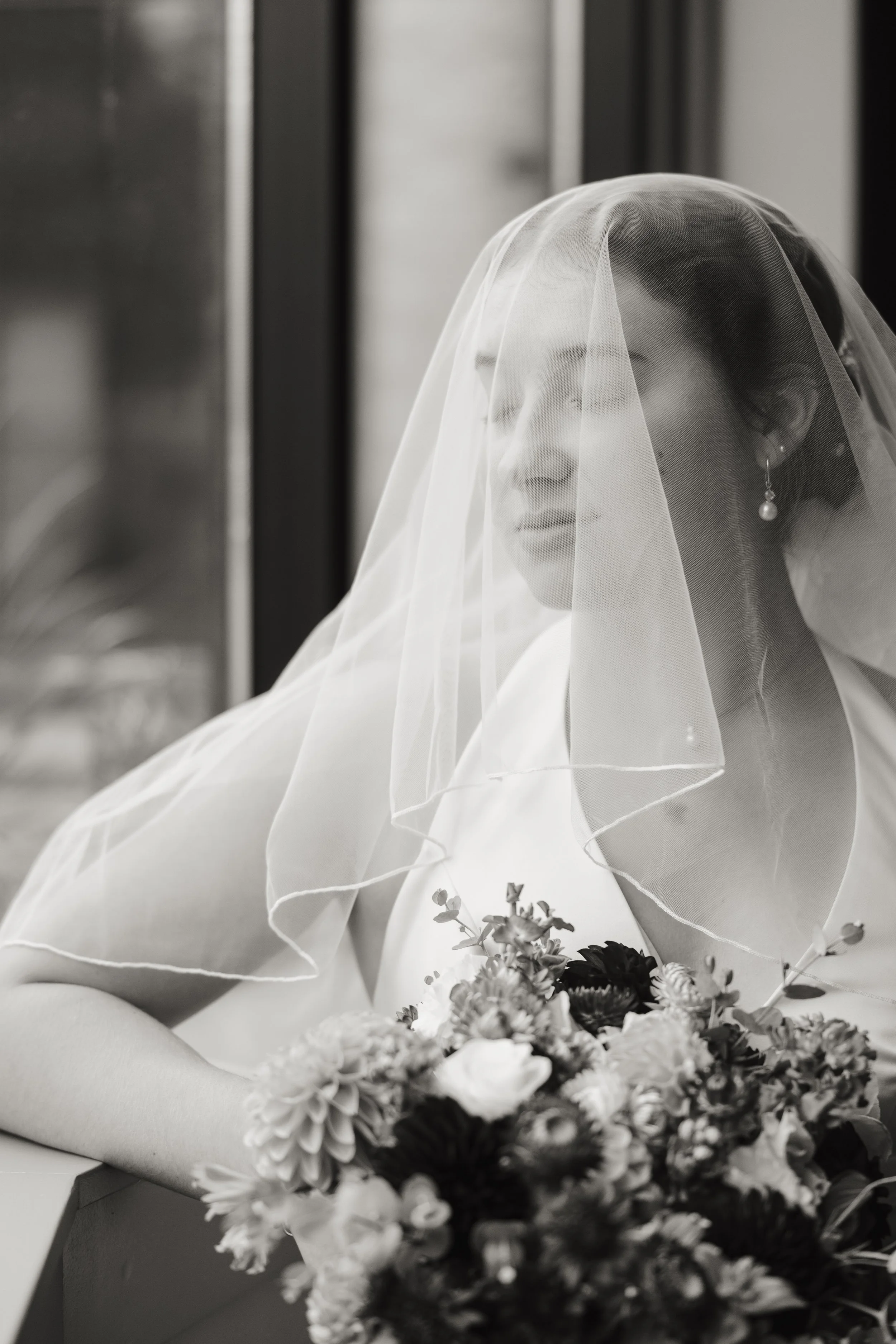 A bride wearing a wedding dress and veil, holding a bouquet of flowers, looking out a window.