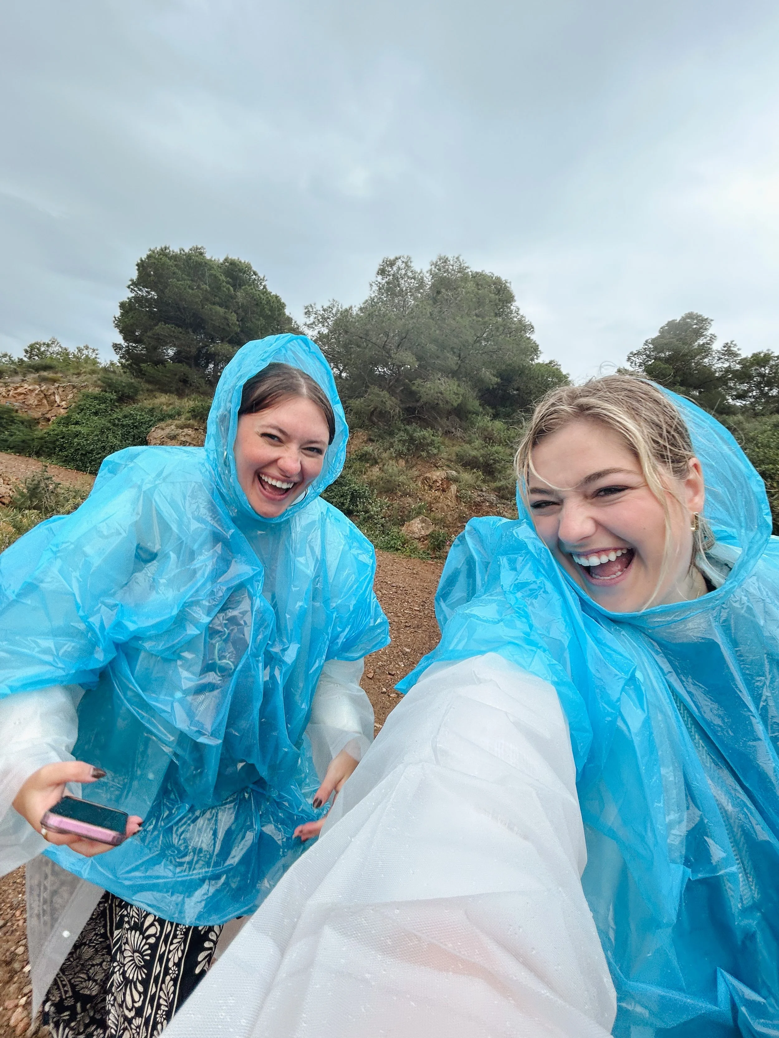 Two women smiling and wearing blue rain ponchos outdoors with trees and cloudy sky in the background.