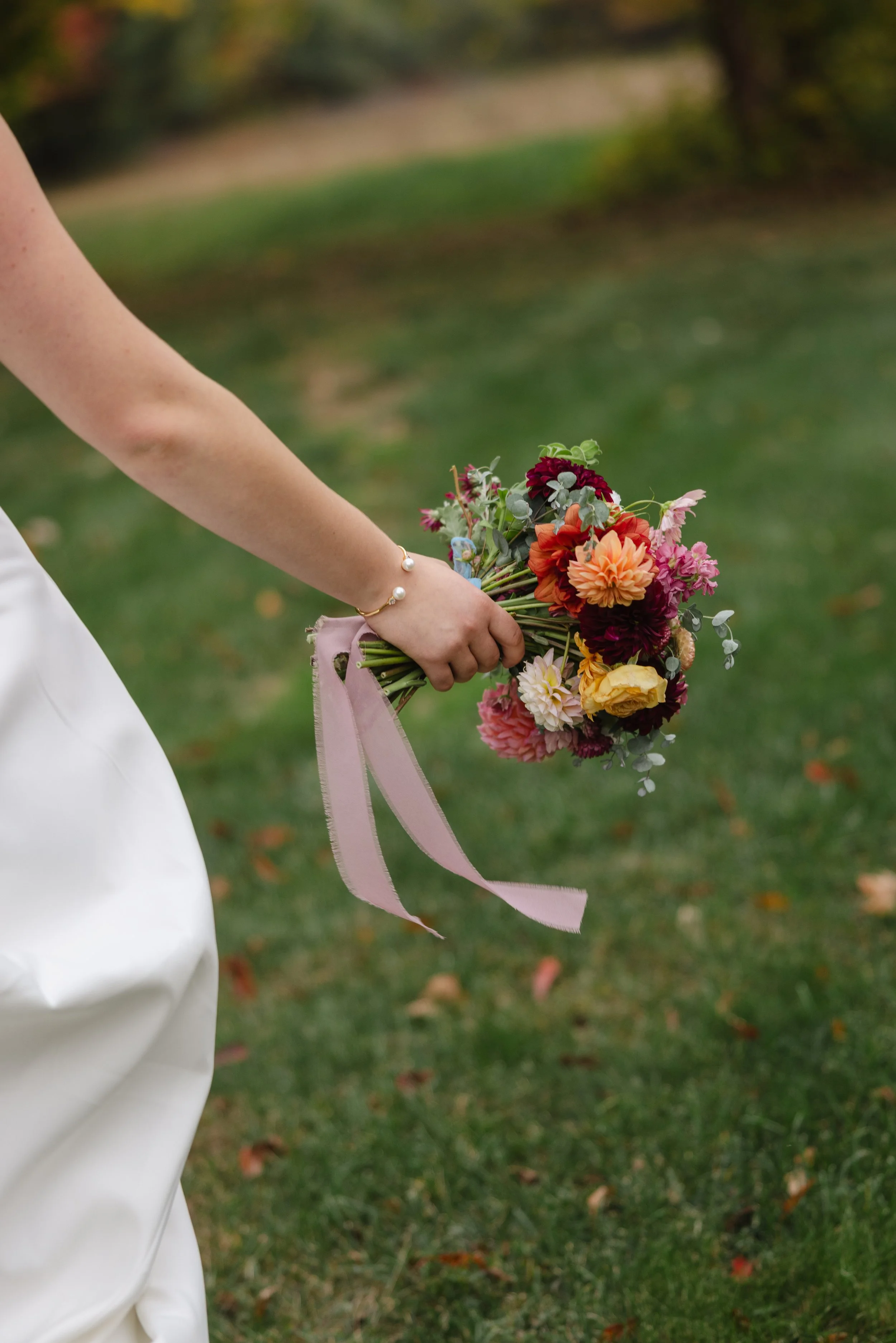 A bride wearing a pearl bracelet on their wrist is holding a colorful bouquet of flowers wrapped with pink ribbon, with a blurred green outdoor background.