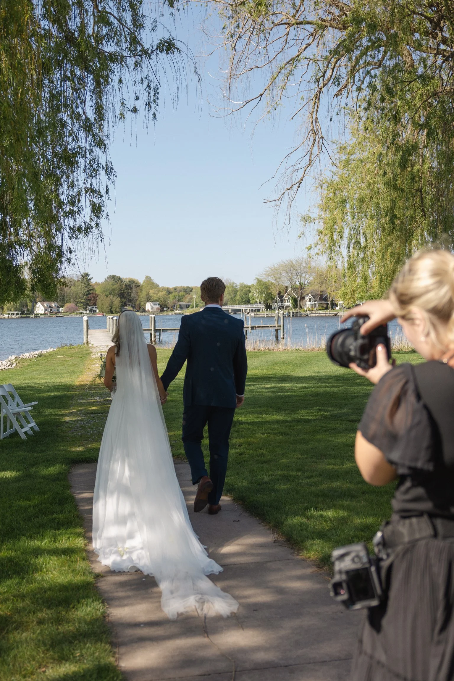A wedding couple walking along a lakeside pathway, with a photographer taking pictures of them amidst trees and blue sky.