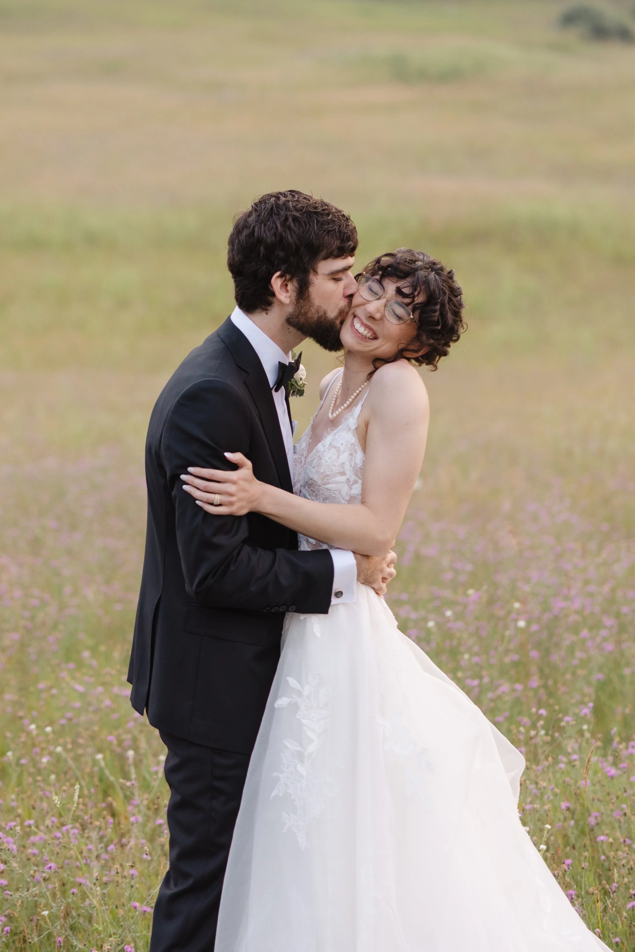A couple in wedding attire embracing in a field of purple flowers, with the groom kissing the bride's cheek as she smiles with closed eyes.