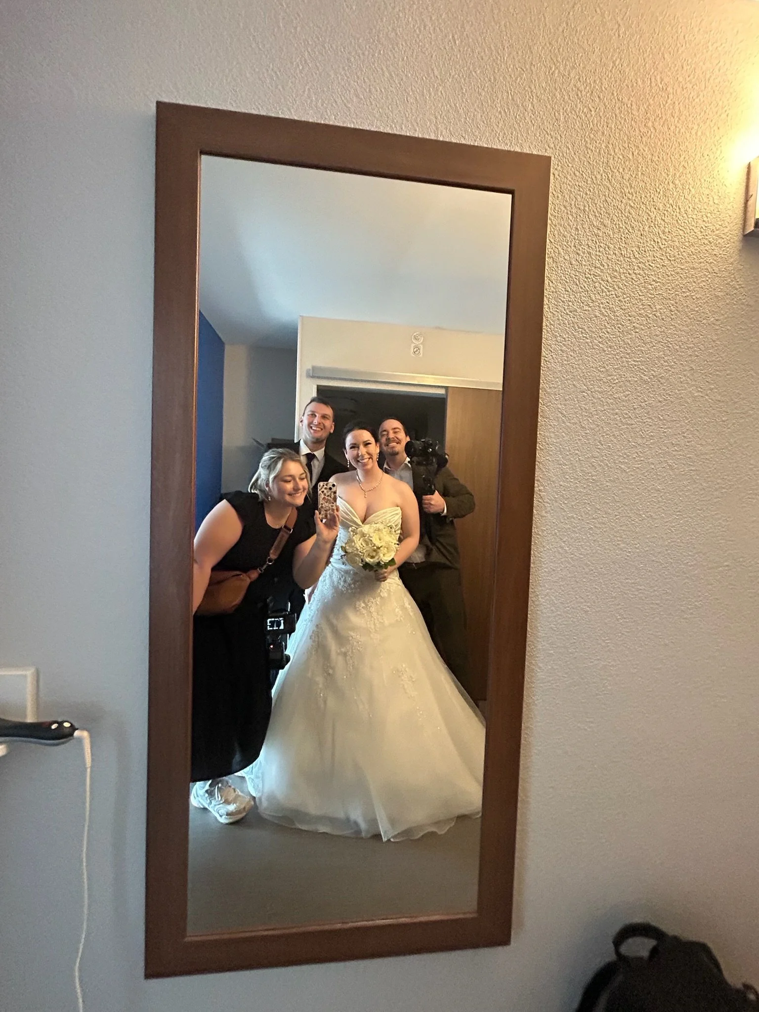 A group of five people, including a bride in a white wedding dress, taking a selfie in front of a mirror in a hotel room. The bride is holding a bouquet of white flowers, and there are smiling friends around her. The room has a wooden-framed mirror a