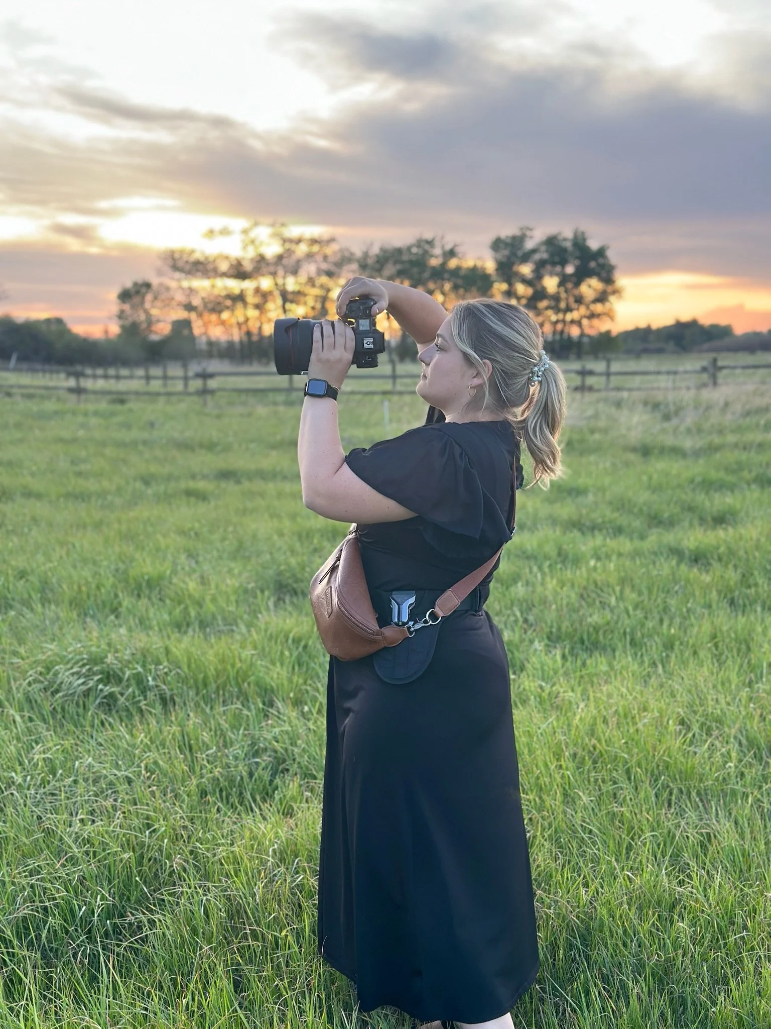 A woman with blonde hair tied in a ponytail, wearing a black top and long black skirt, stands in a green grassy field at sunset, taking a photograph with a camera.