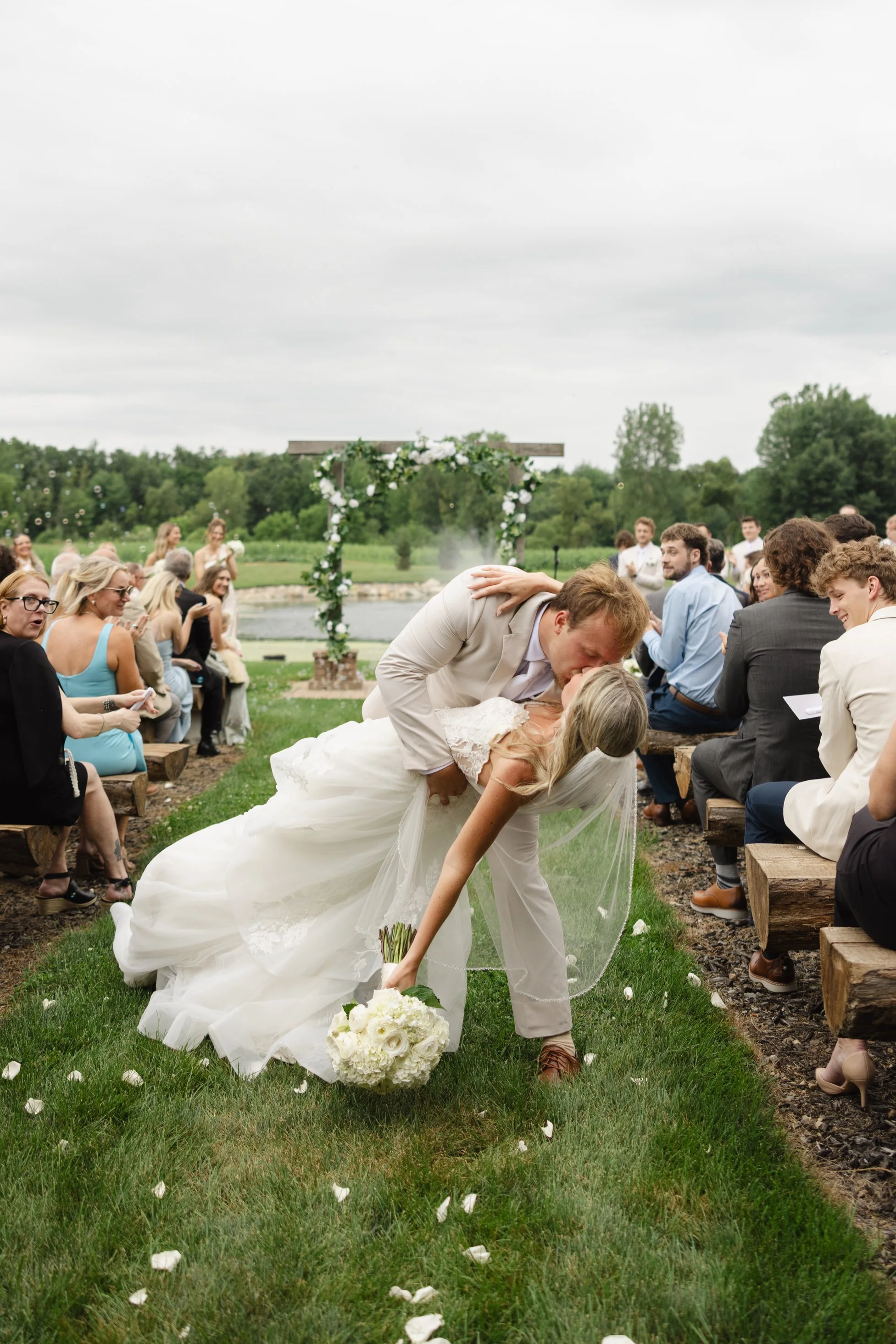 A wedding ceremony outdoors by a lake featuring a bride and groom sharing a kiss while leaning over a bouquet, with guests seated on wooden benches on either side, and a decorated arch in the background. The sky is overcast.