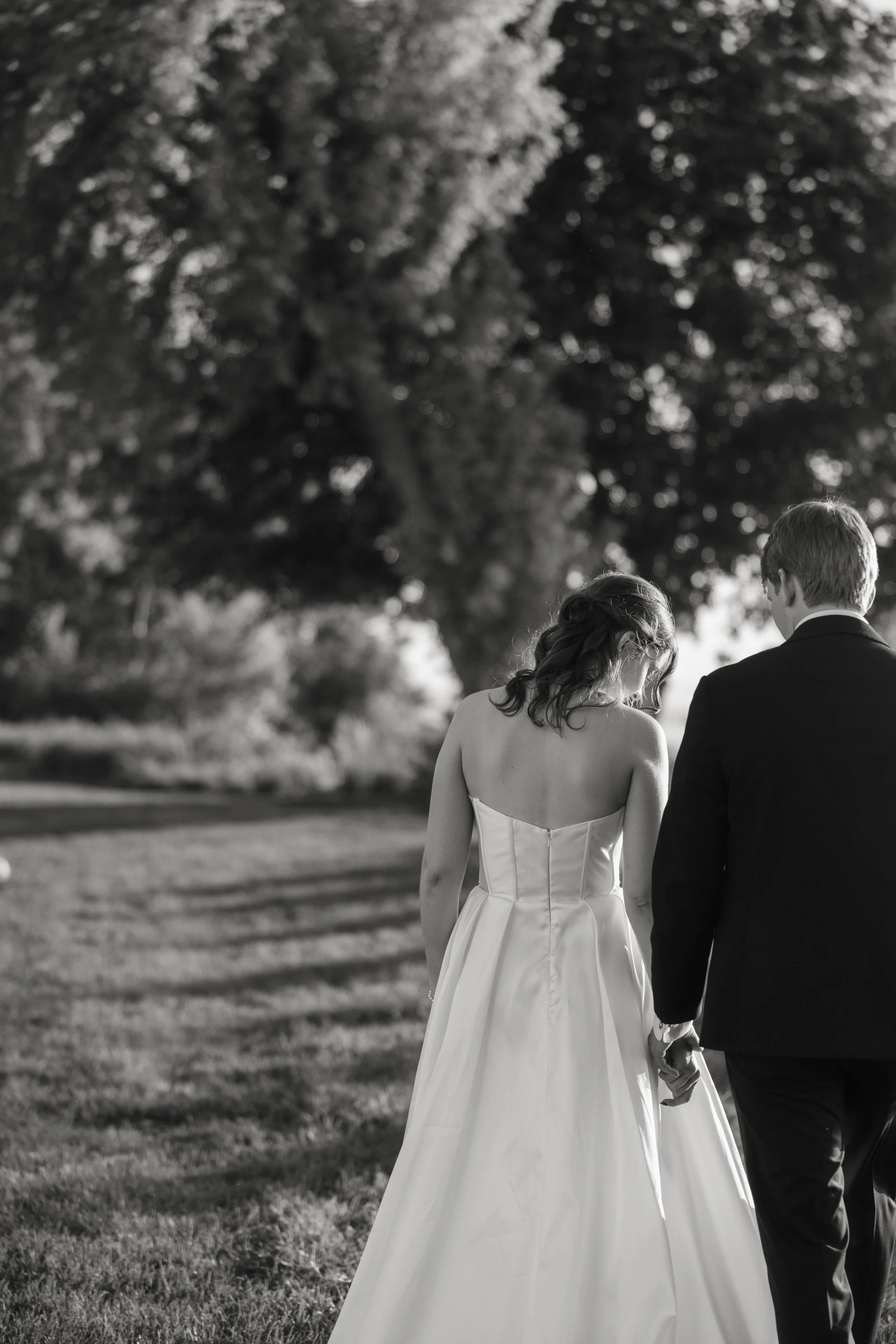 A black and white photo of a bride and groom holding hands, walking outdoors in a park or garden, with large trees in the background.