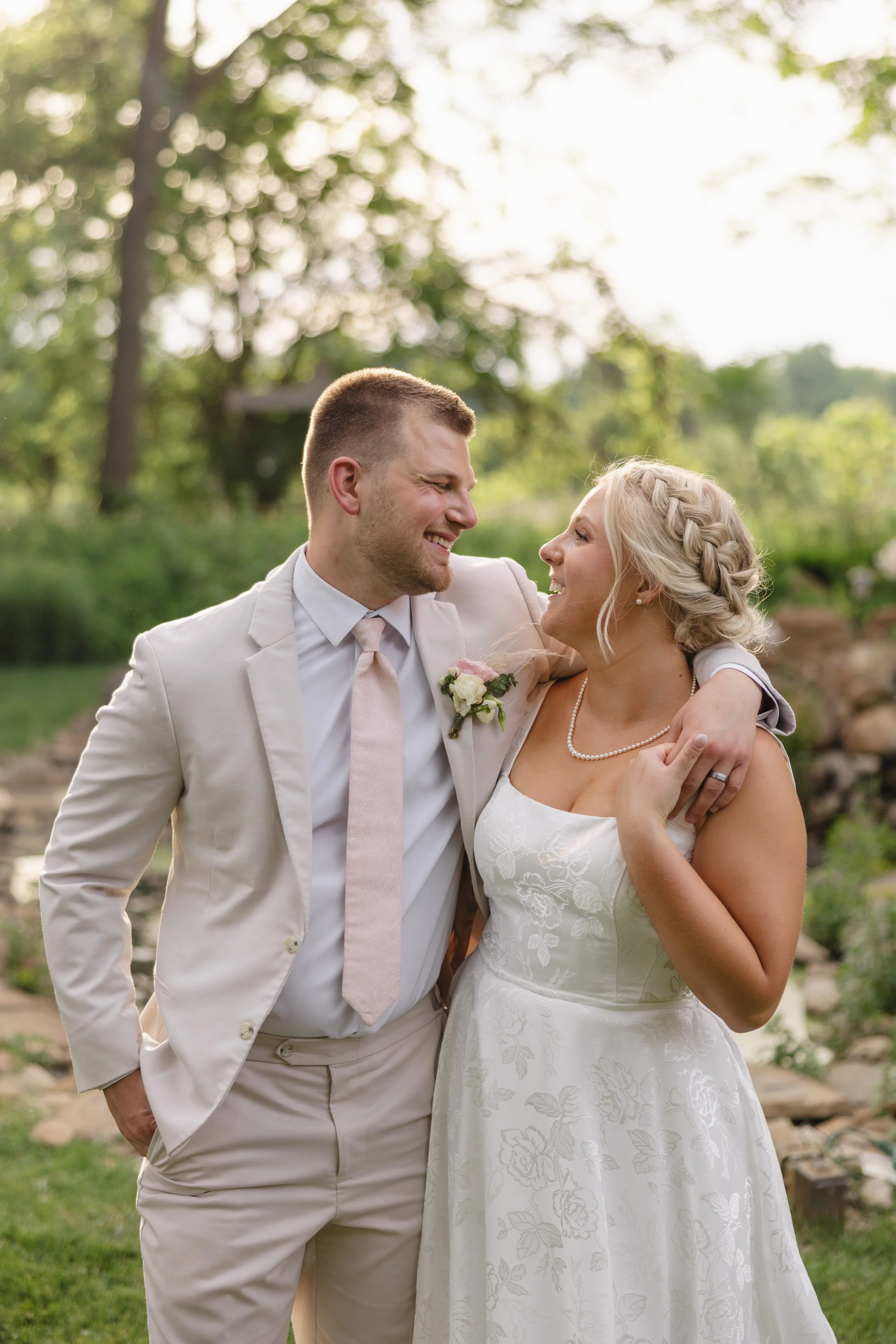 A newlywed couple smiling and looking at each other outdoors during the day, surrounded by green trees and grass.