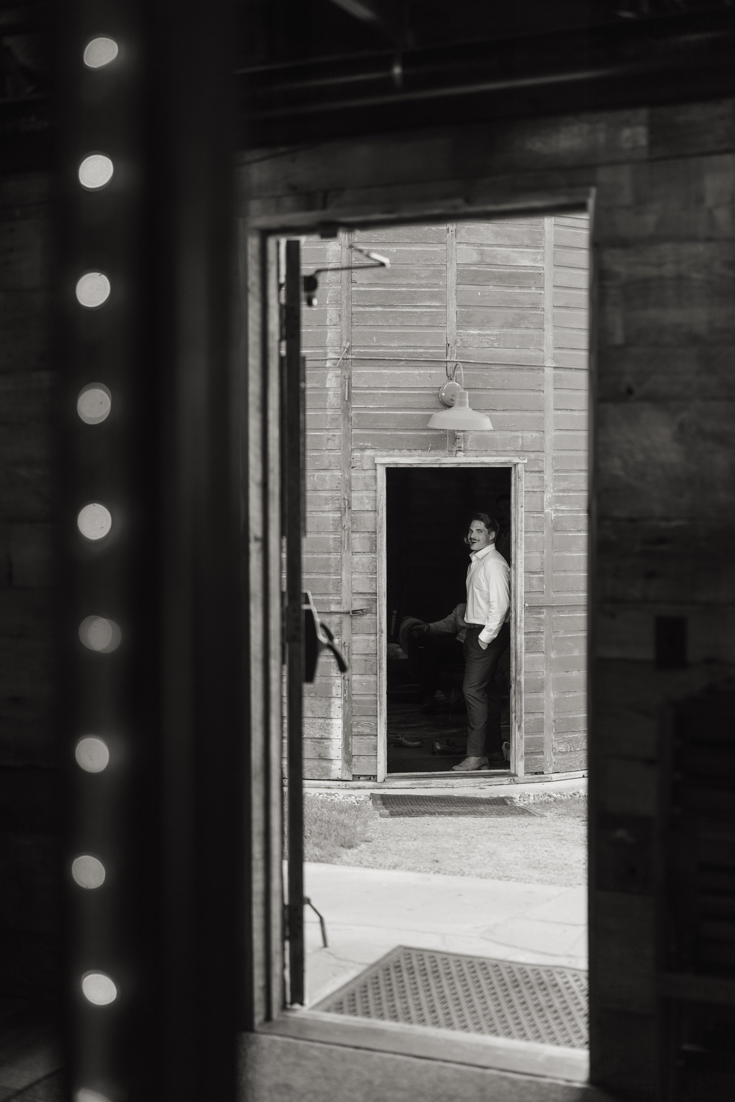 A man in a white shirt and dark pants standing and smiling outside a wooden building, viewed through the doorway of a dimly lit room.