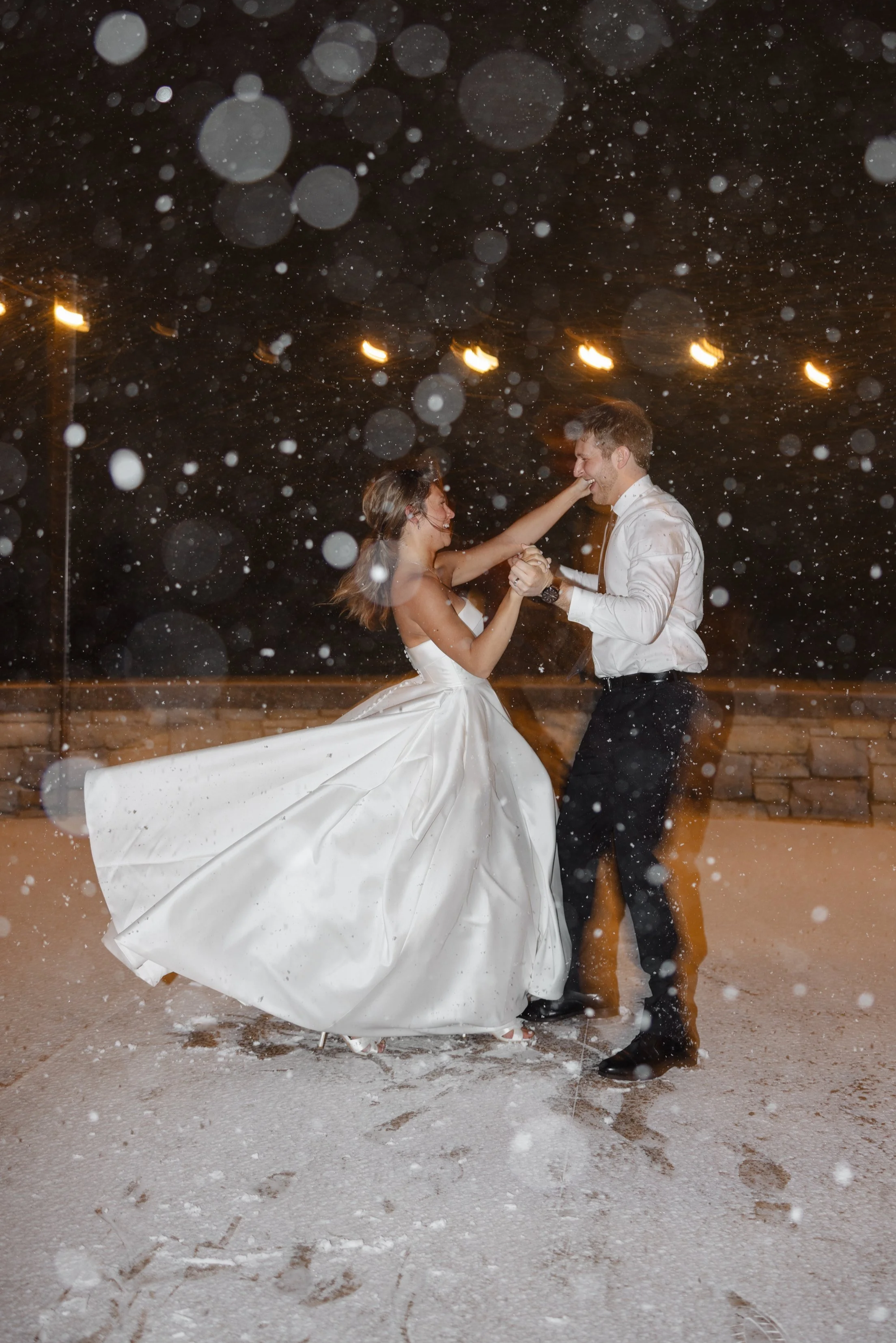 A bride and groom dancing outside at night with snow falling around them, illuminated by string lights overhead.