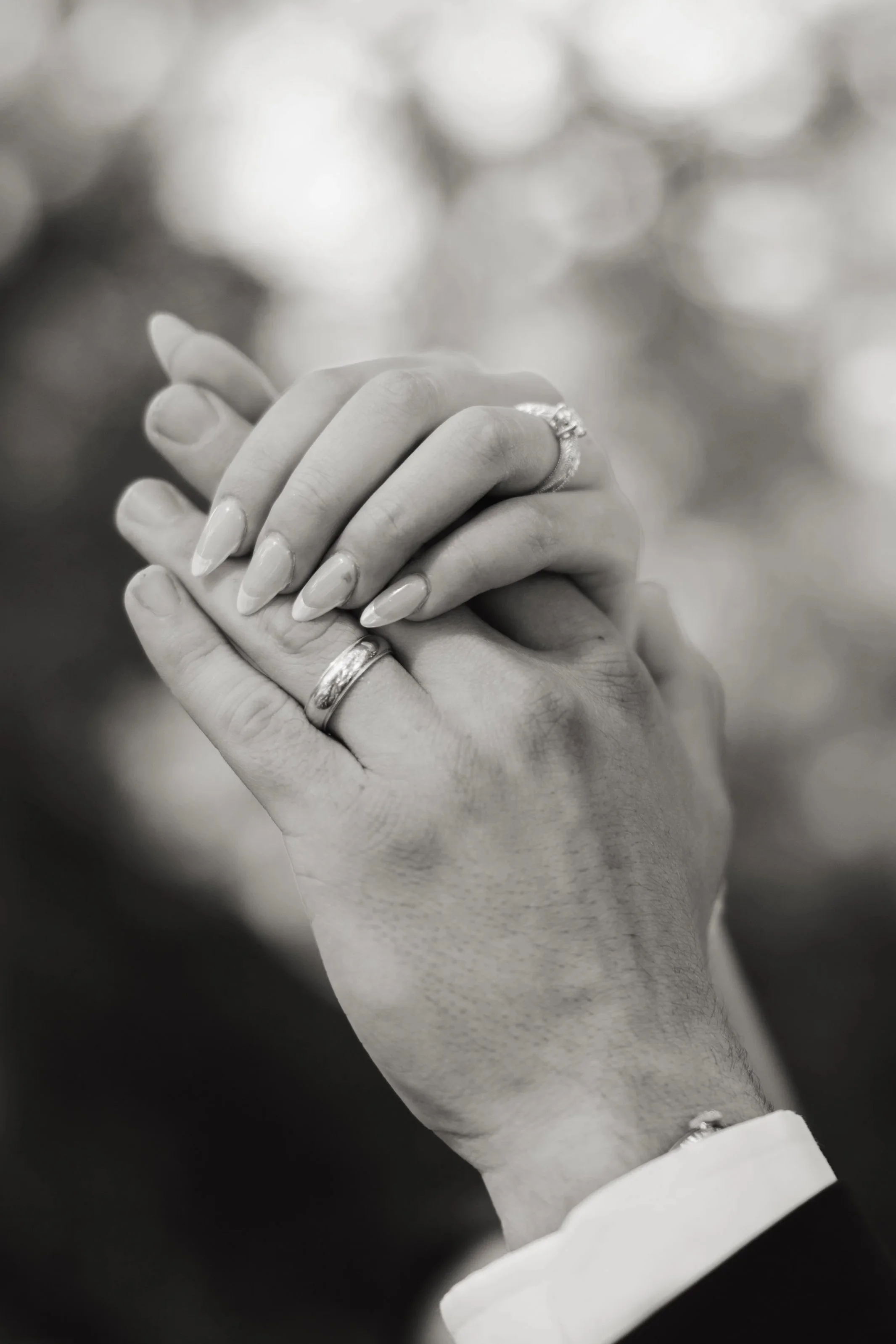 A close-up black and white photo of two hands, with fingers interlaced, displaying wedding rings. The person is wearing a white cuffed shirt.