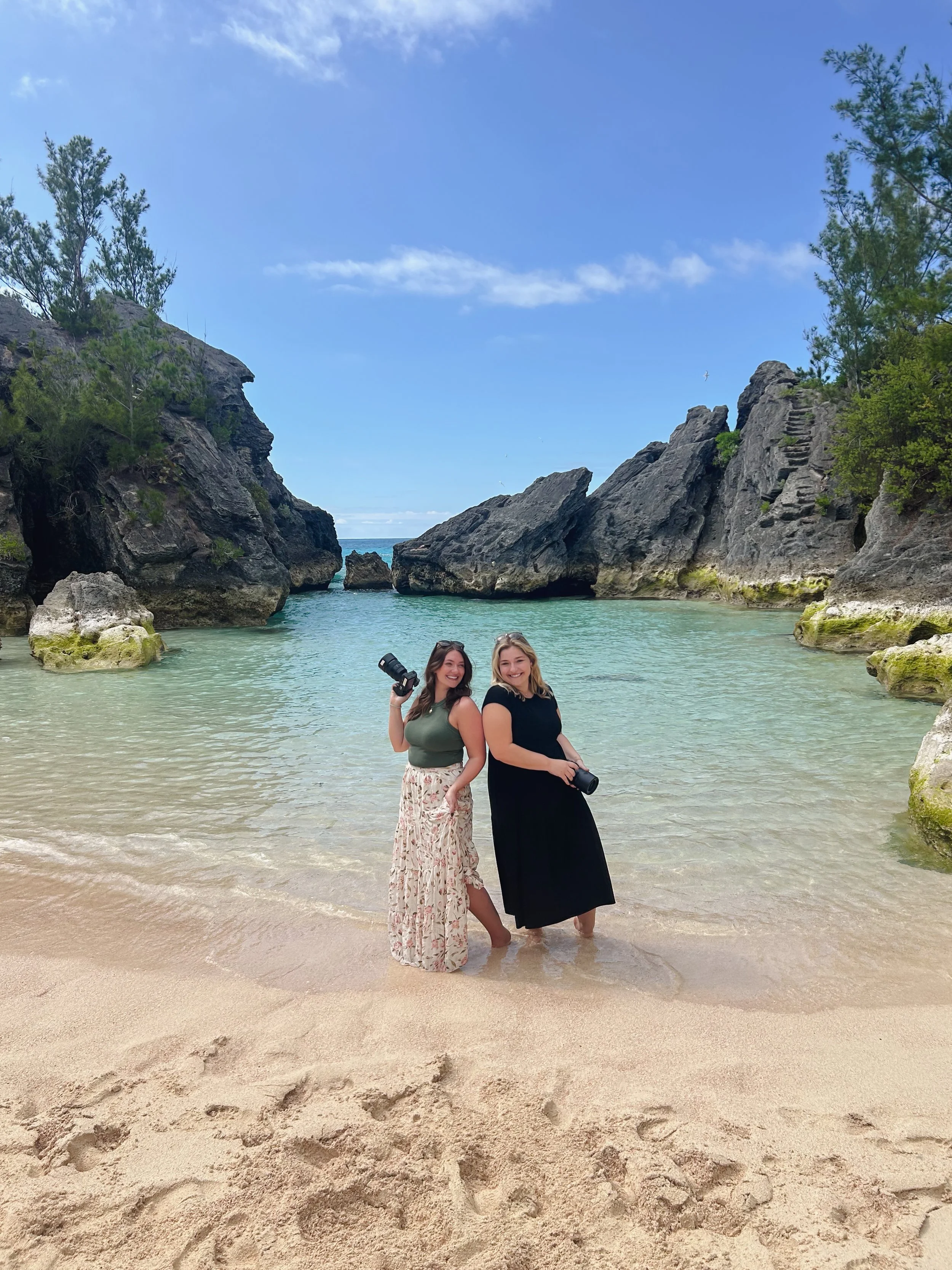 Two women standing in shallow water on a beach with rocky cliffs and green trees in the background.