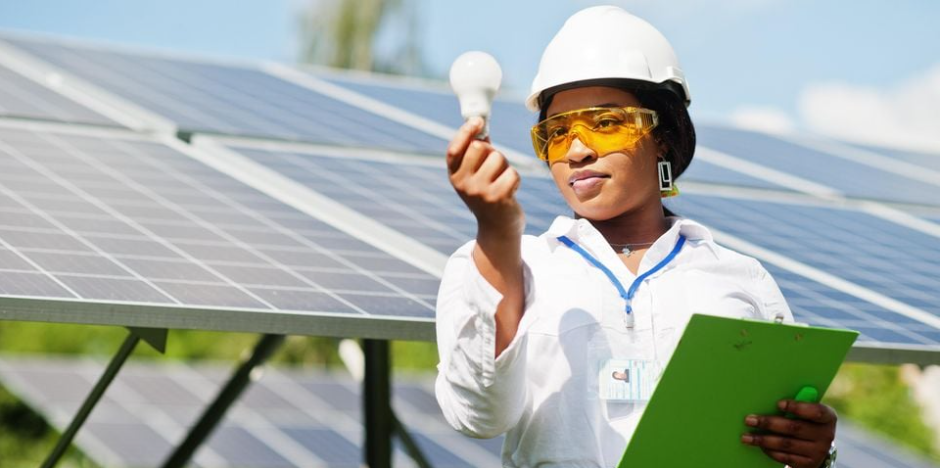 A woman wearing a white safety helmet and yellow safety glasses examines a solar light bulb while holding a green clipboard at a solar panel farm.