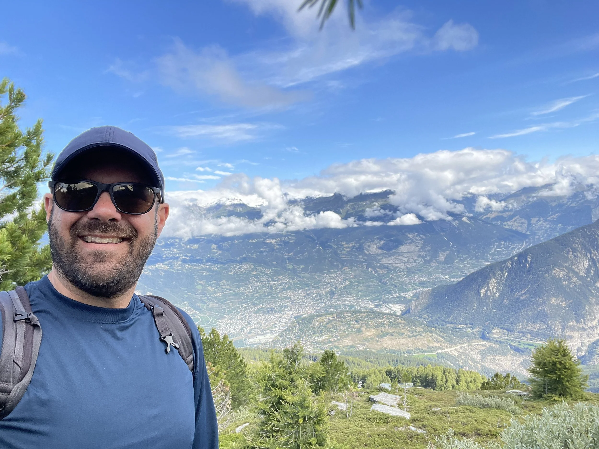 Dan Weber on a mountain trail, overlooking a valley with mountains, trees, and clouds in the background.