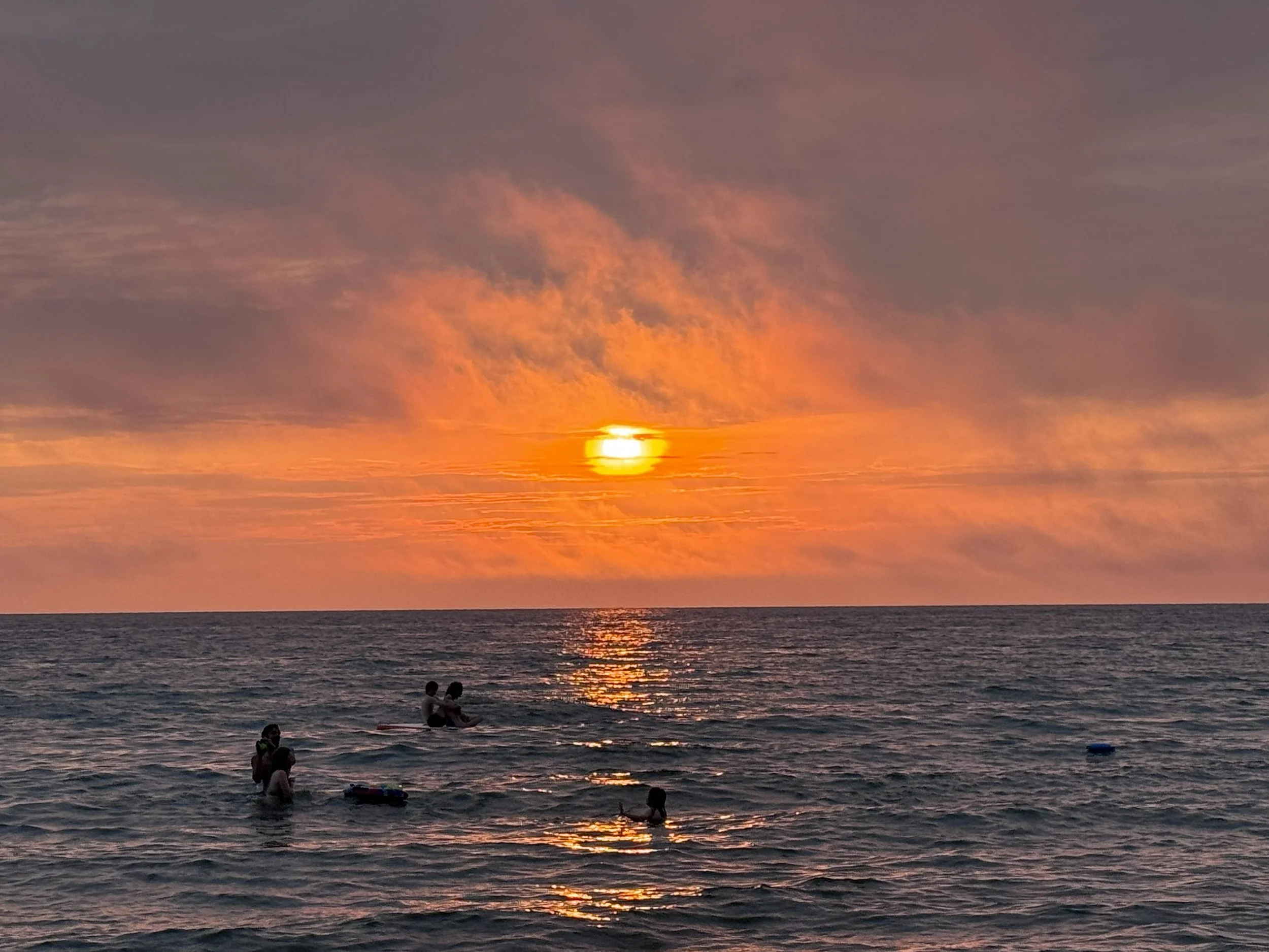 A sunset over Lake Michigan with pink, orange, and purple clouds. People are swimming and relaxing in the water, some sitting on surfboards, enjoying the tranquil scene.