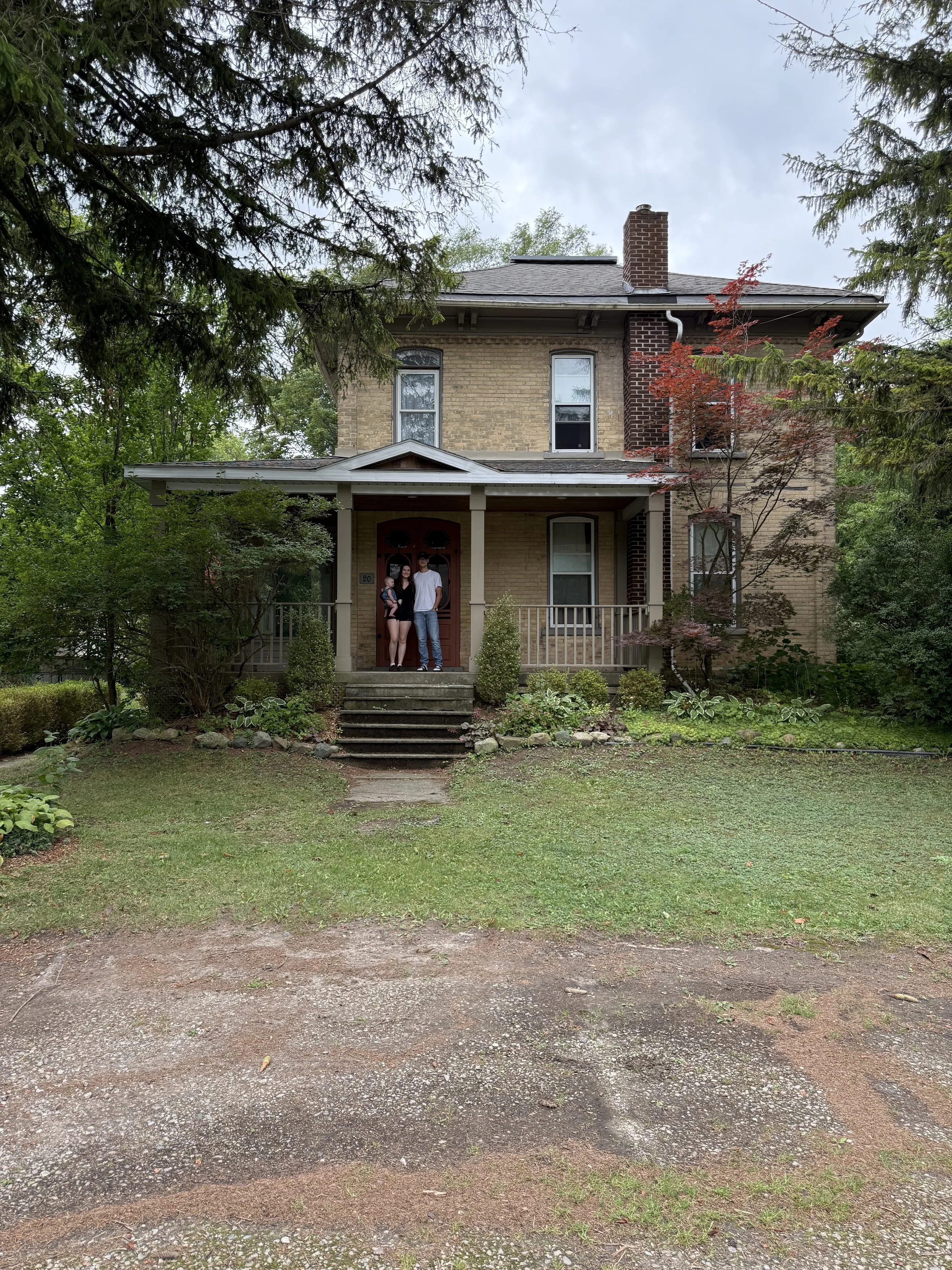 A two-story house with a brick exterior and a front porch. A family of three is standing on the porch, surrounded by trees and greenery.