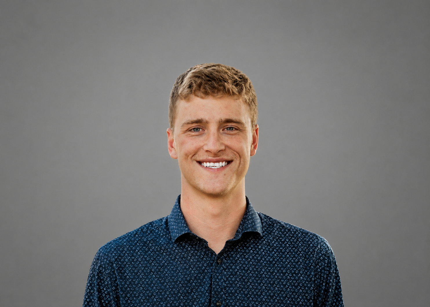 A young man with short, wavy blonde hair and blue eyes smiling at the camera against a plain gray background, wearing a navy blue button-up shirt with a small pattern.