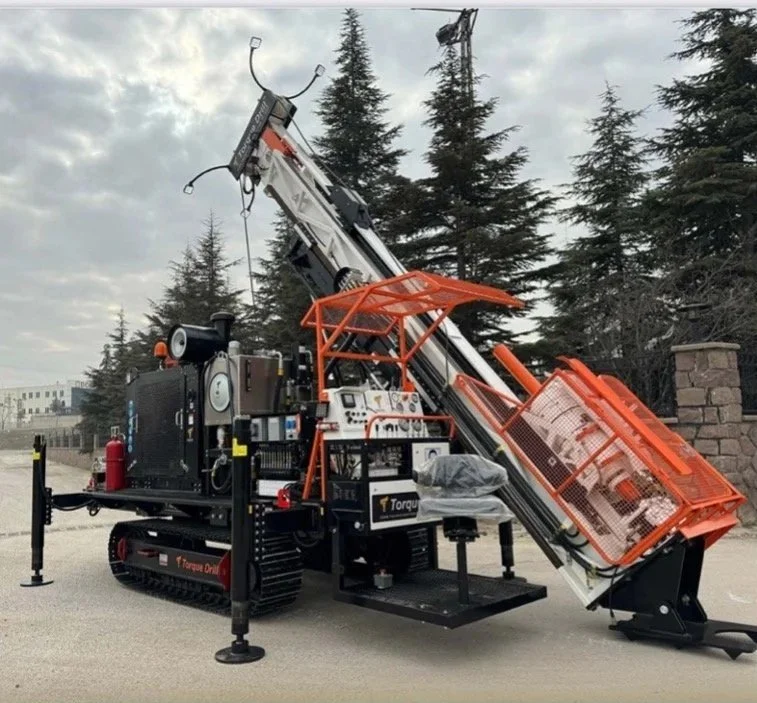 A large drilling machine with tracked wheels and an orange safety cage, positioned outdoors on a paved area with trees and a stone wall in the background.