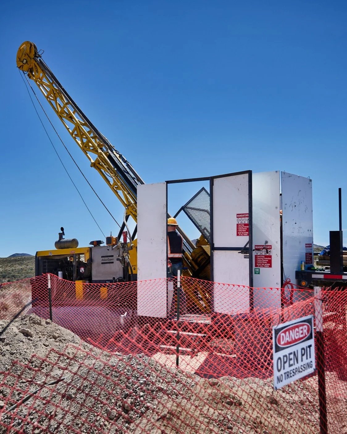 Construction site with a crane and a worker inside a fenced area under a clear blue sky. Warning signs read 'Danger Open Pit No Trespassing.'