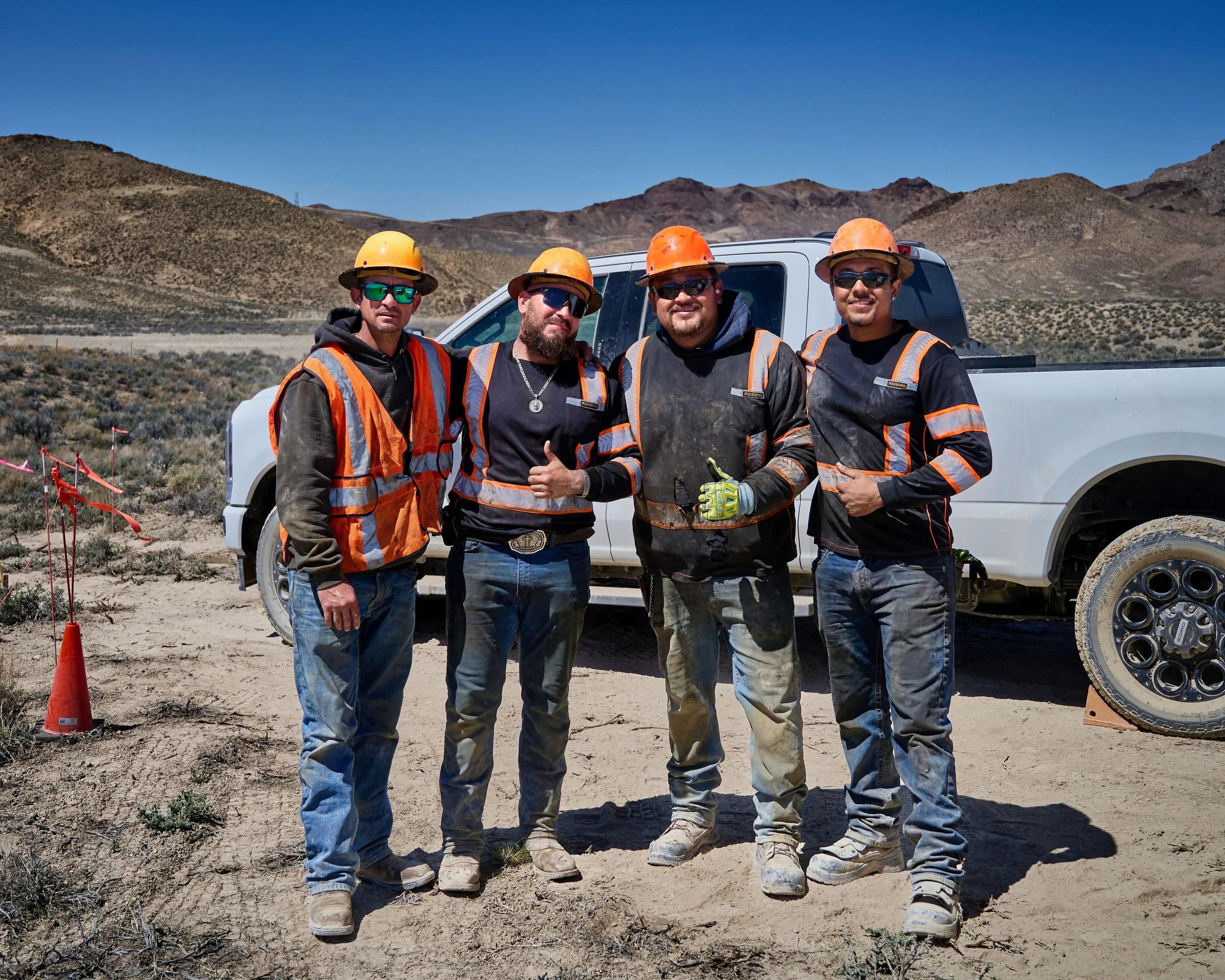 Four construction workers wearing orange safety helmets and reflective vests standing outdoors in front of a white pickup truck with mountains in the background under a clear blue sky.