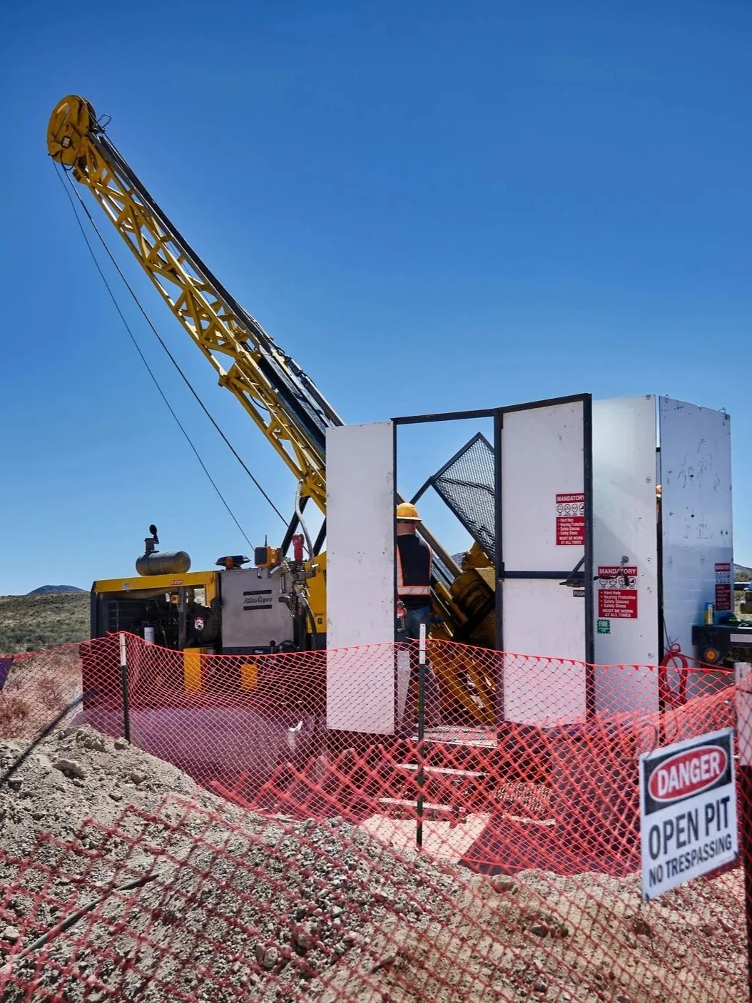 A construction site with a large yellow tower crane and a worker wearing a helmet operating some equipment inside a small, enclosed white office space. There is orange safety netting around the site and a sign that says 'Danger Open Pit No Trespassing'.