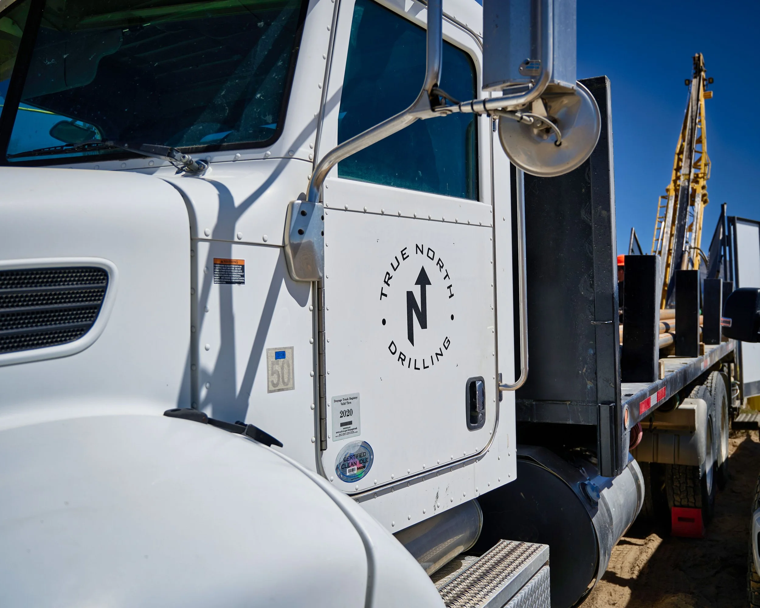 Close-up of a white industrial truck with a logo and stickers on the door, a side mirror, and a chrome handrail, with yellow construction equipment in the background against a clear blue sky.