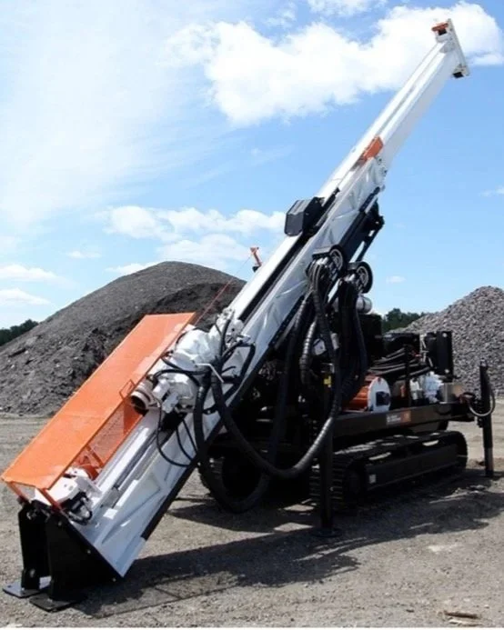 A large industrial drilling machine operated outdoors, with a long, inclined drill pipe, on a construction or mining site with gravel piles in the background.