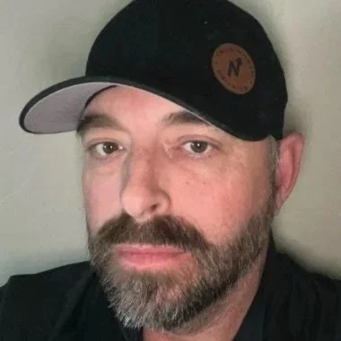 A man with a beard wearing a black cap with a leather patch, looking at the camera indoors.