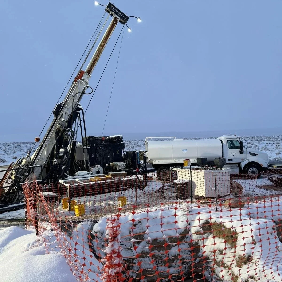 A drilling rig and a water truck at a snowy construction or pipeline site surrounded by orange snow fencing.