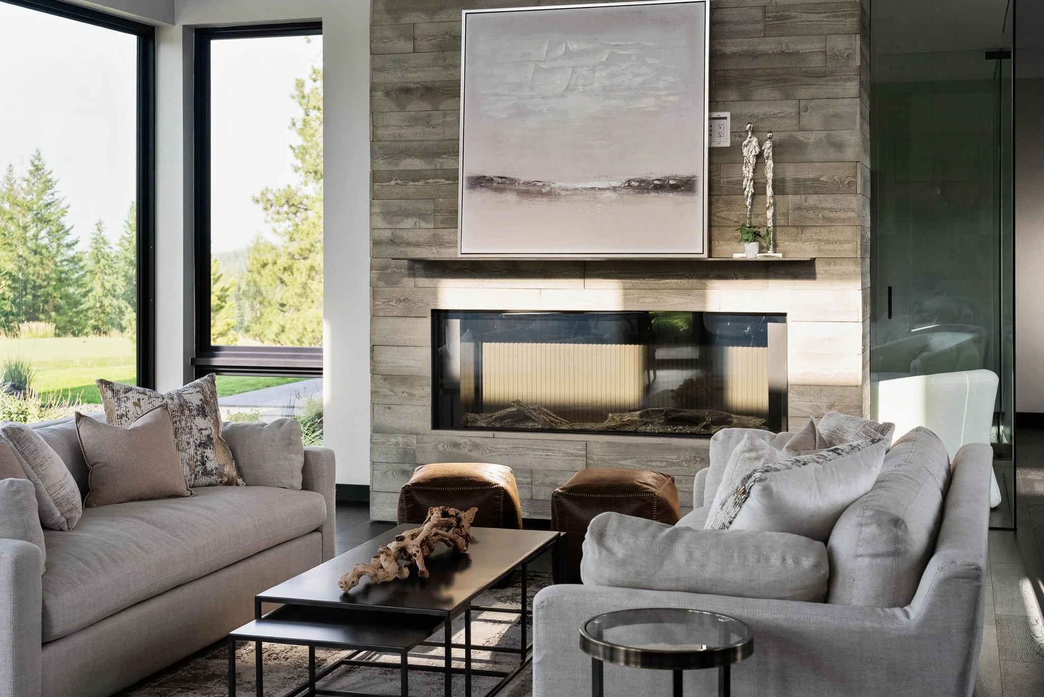 Modern living room staged by Home Staging Works featuring neutral sofas, a stone‑clad fireplace and floor‑to‑ceiling windows framing a forest view, illustrating the transformation achieved through staging.