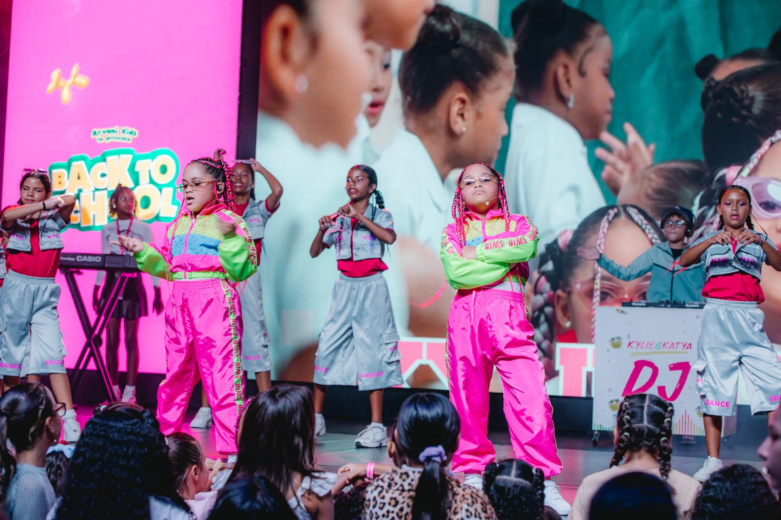 Young girls performing a dance on stage during a school event, with an audience of children watching from below. The stage background has a pink sign that says 'Back to School' and other colorful decor.