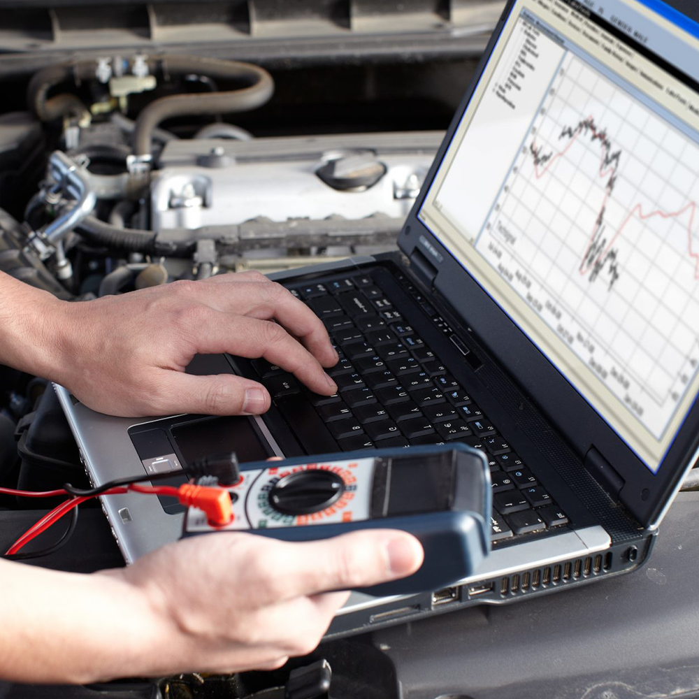Person using a laptop with a graph on the screen to diagnose car engine problems, holding a multimeter.