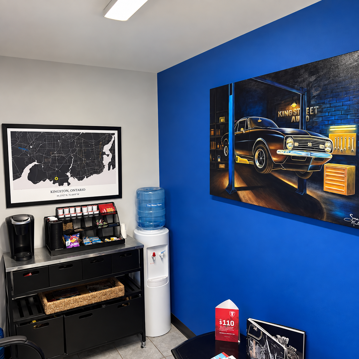 Interior of a shop or office with a blue accent wall, artwork of a classic black car in a garage setting, a map of Kingston, Ontario, a water cooler, a coffee station, and promotional materials on a table.