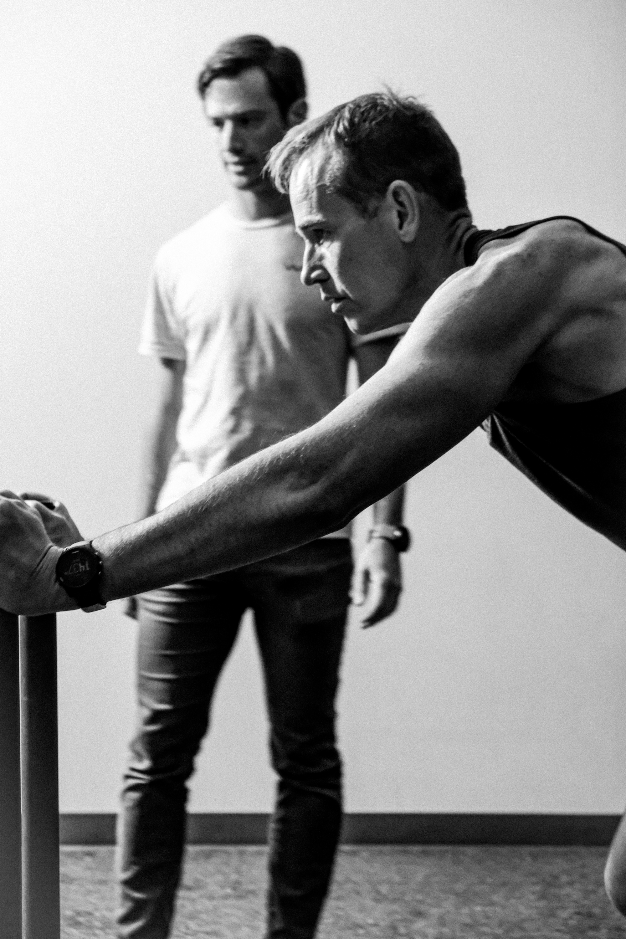 Two men with athletic builds, one with light-colored hair and the other with darker hair, are engaged in an activity together. The man with dark hair is leaning forward and focusing intently, while the other man stands in the background observing. The setting appears to be indoors with a plain wall and carpeted floor.