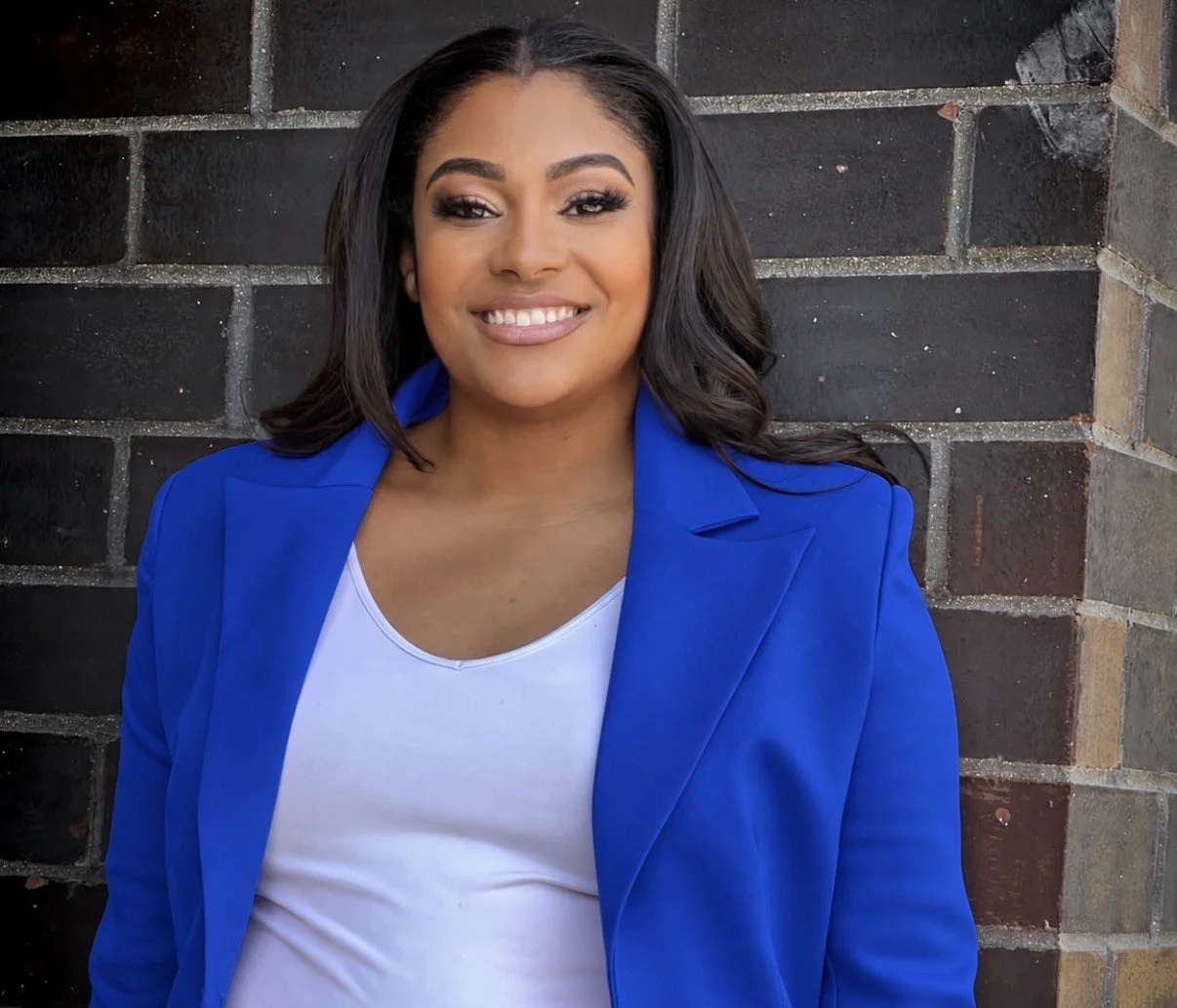 Smiling woman with dark hair wearing a bright blue blazer and white top, standing against a dark brick wall.