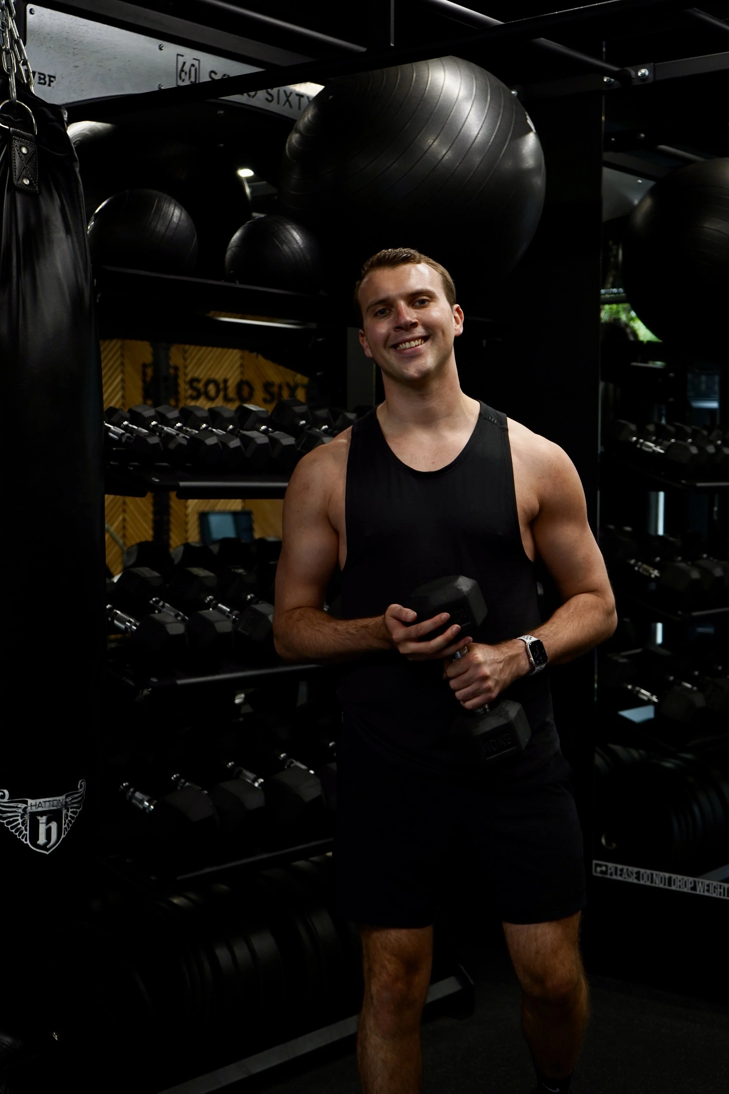 A smiling man in a black tank top and shorts holding a dumbbell at a gym with black workout equipment and black exercise balls in the background.