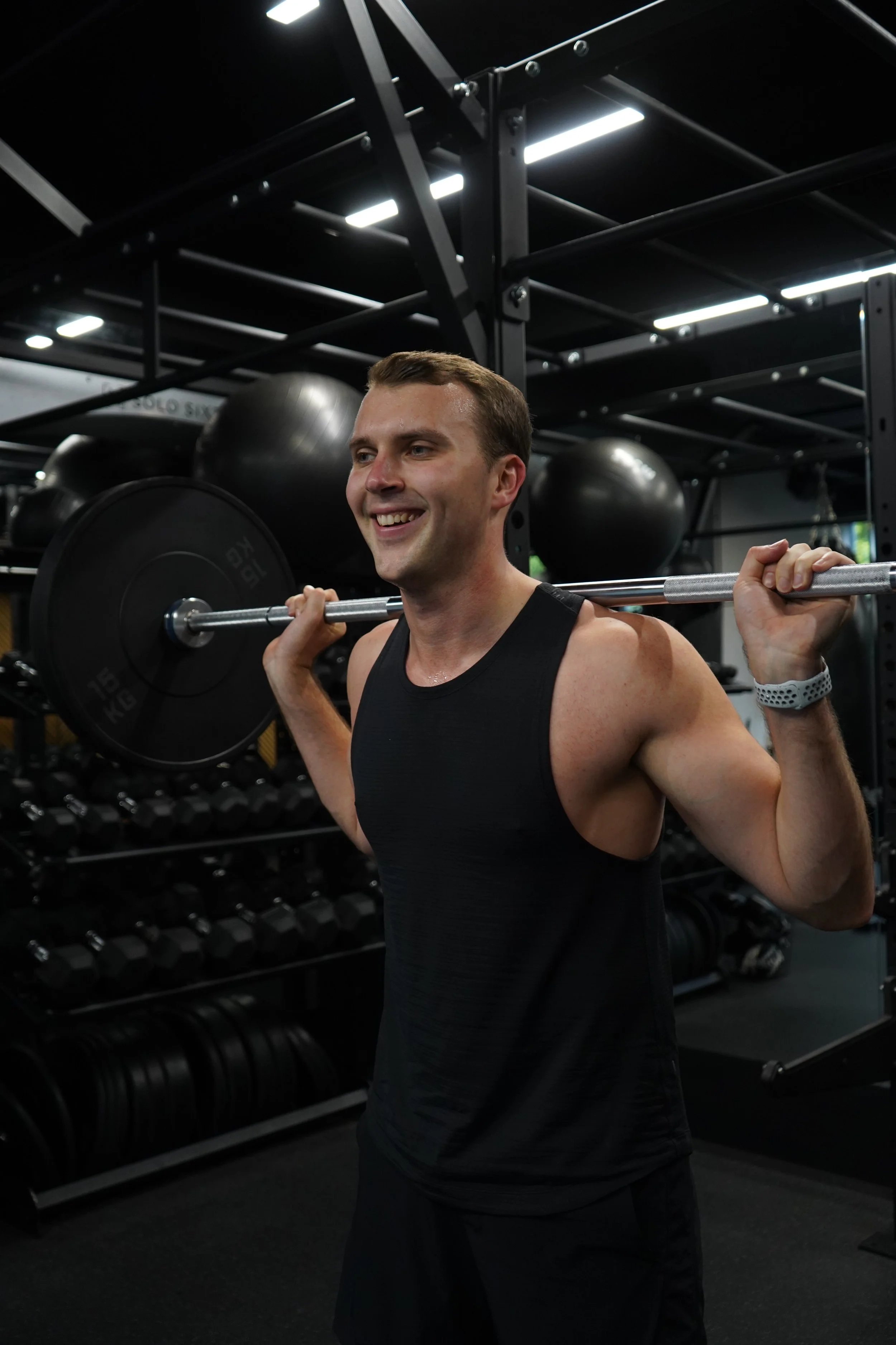 A man smiling and lifting a barbell on his shoulders in a gym with black weights and equipment in the background.