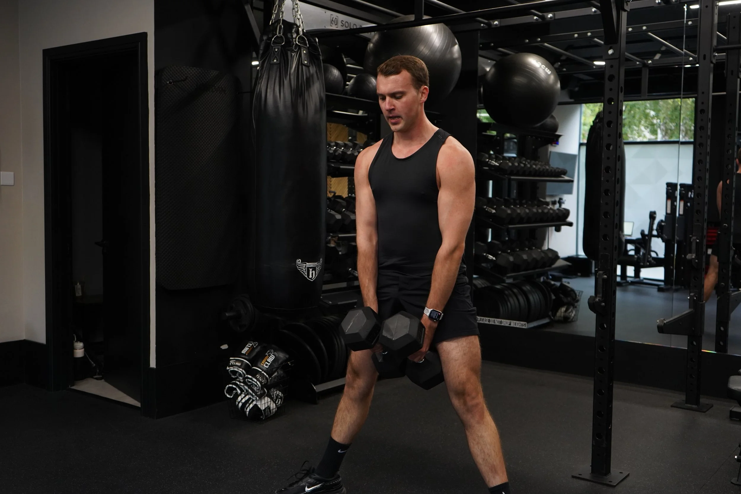 A man in black athletic clothing lifting dumbbells in a gym with black equipment and a punching bag.