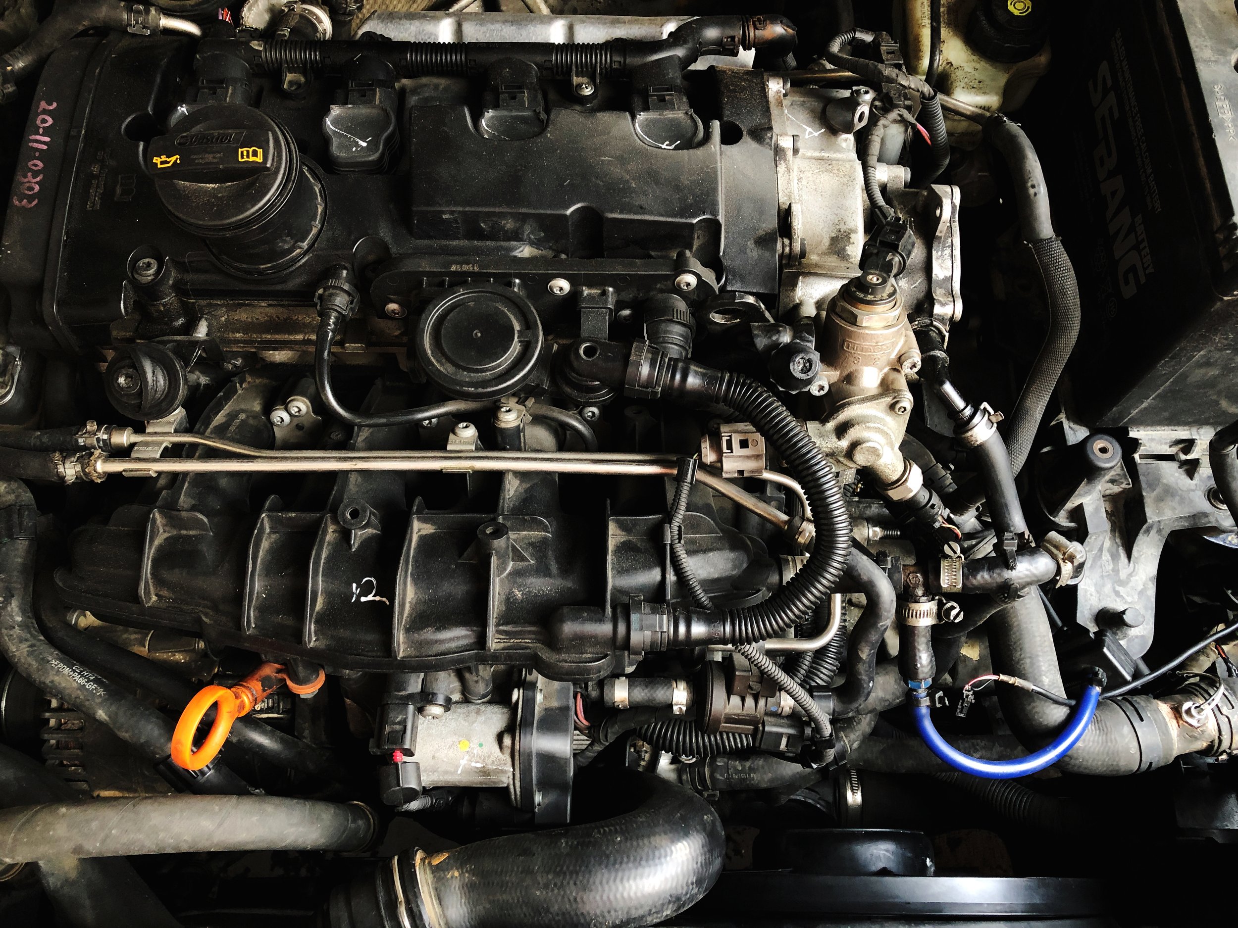 A detailed view of a car engine with various black hoses, wires, and metal components. The image shows parts such as the oil cap, radiator hoses, and other engine parts, with a slightly dusty appearance.