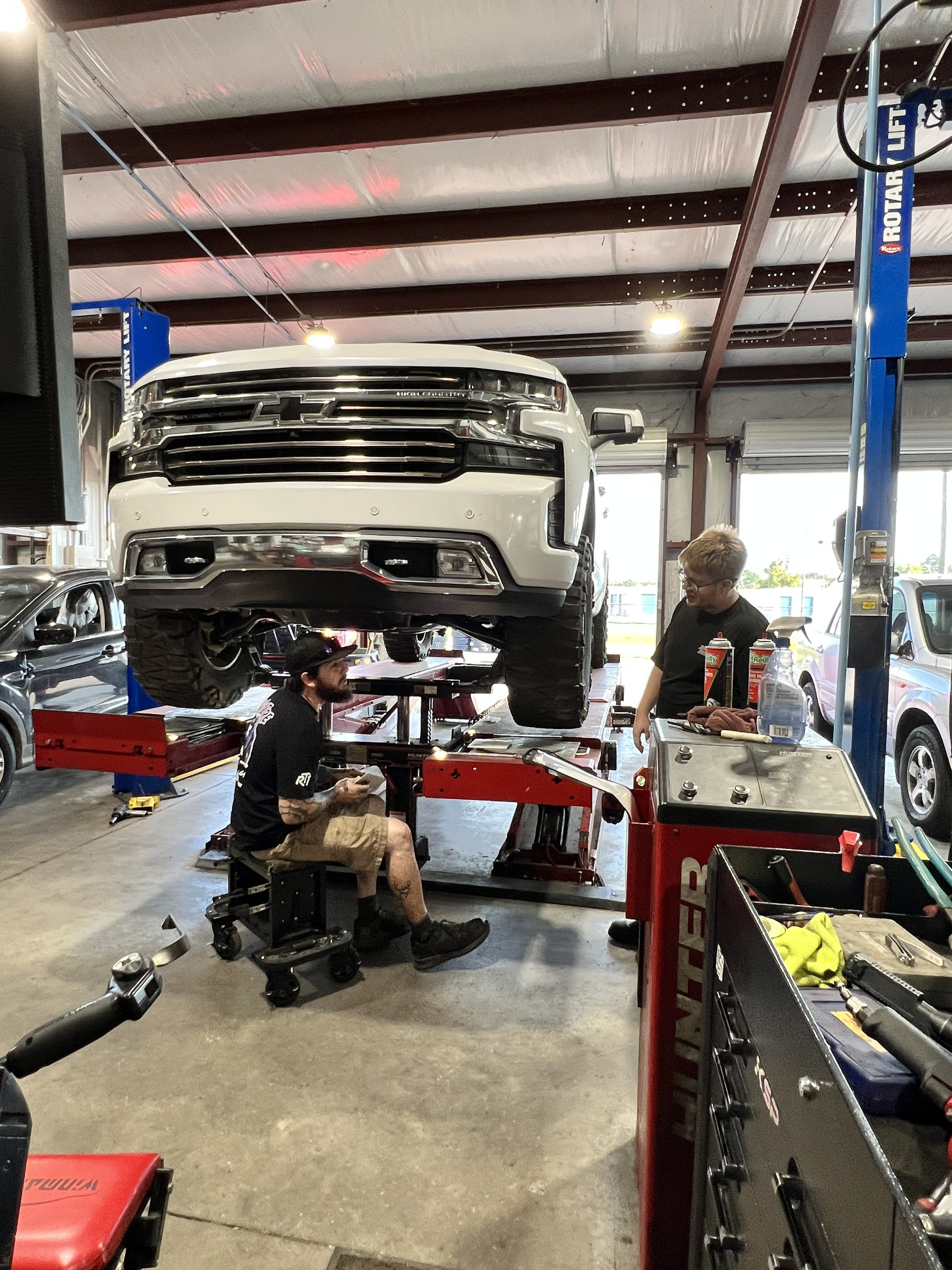 Two mechanics working on a white pickup truck lifted on a hydraulic lift inside an auto repair shop. One mechanic is sitting at a workbench, and another is standing nearby. The shop has large windows and various tools and equipment around.