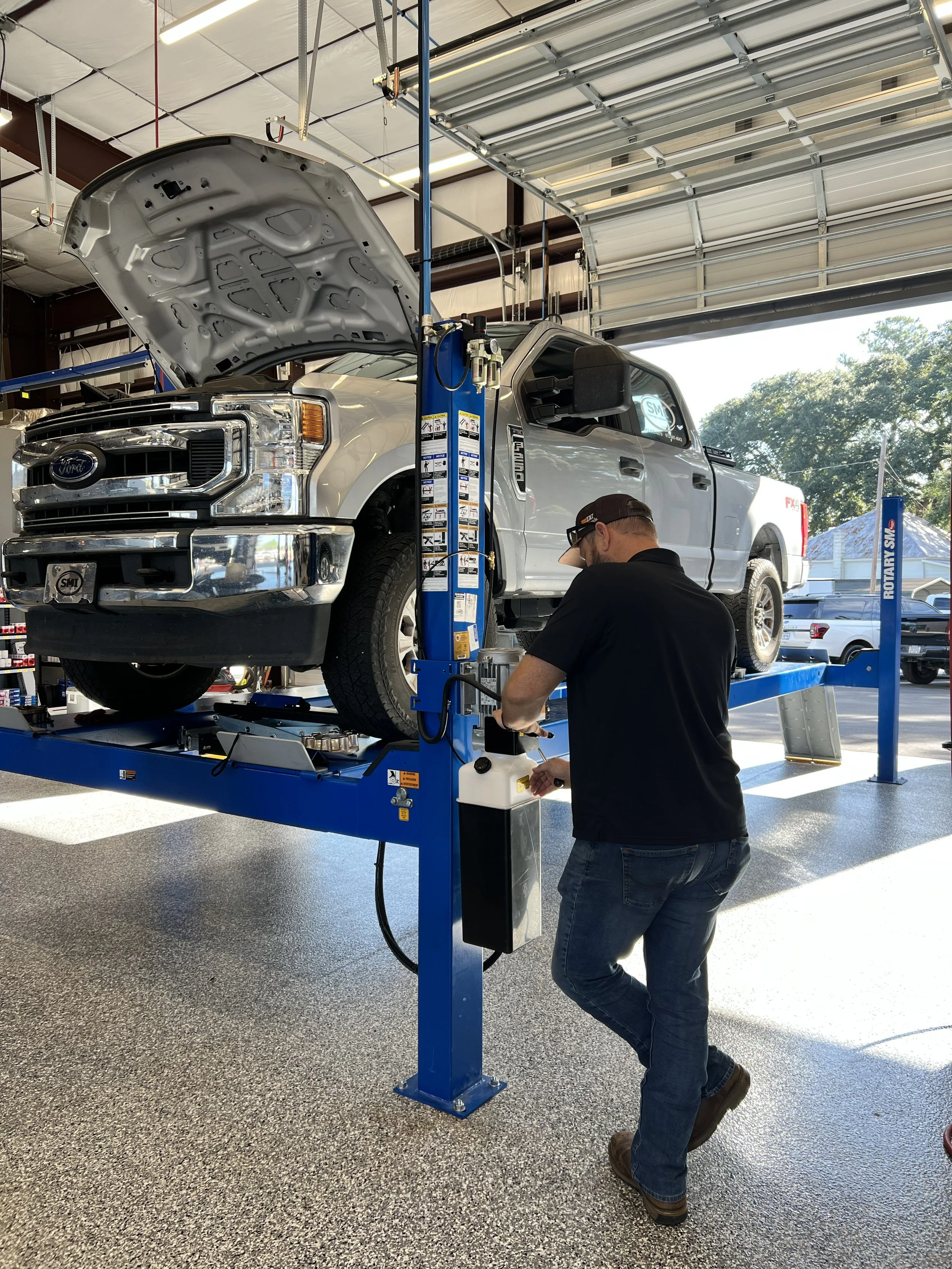 A mechanic working on a white Ford truck elevated on a blue hydraulic lift inside an auto repair shop.