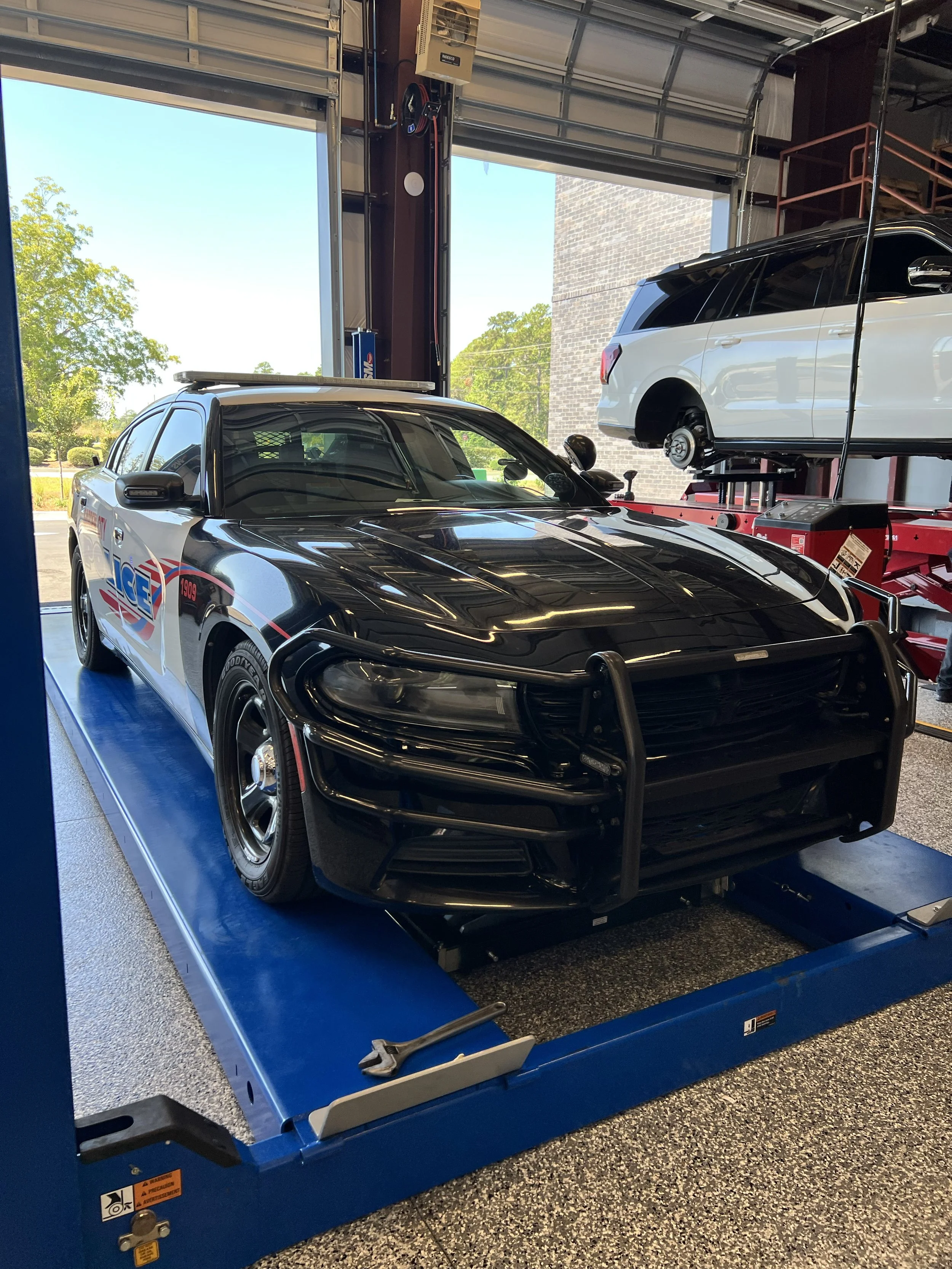 A black police car with police markings and a push bumper is inside a garage on a lift, with a white vehicle on a hydraulic lift in the background.