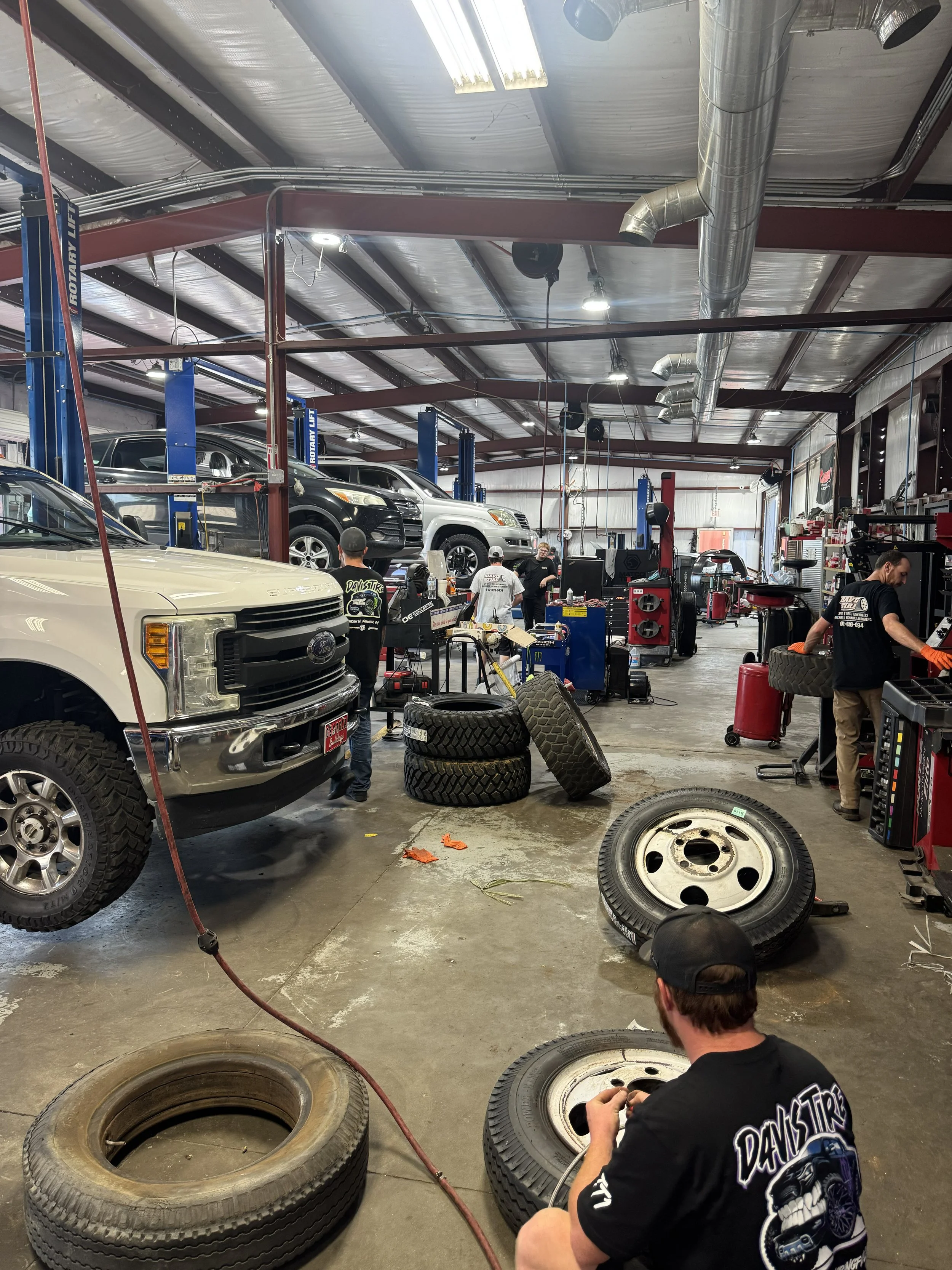 Mechanics working in an automotive repair shop with vehicles on lifts, tires, and tools around.