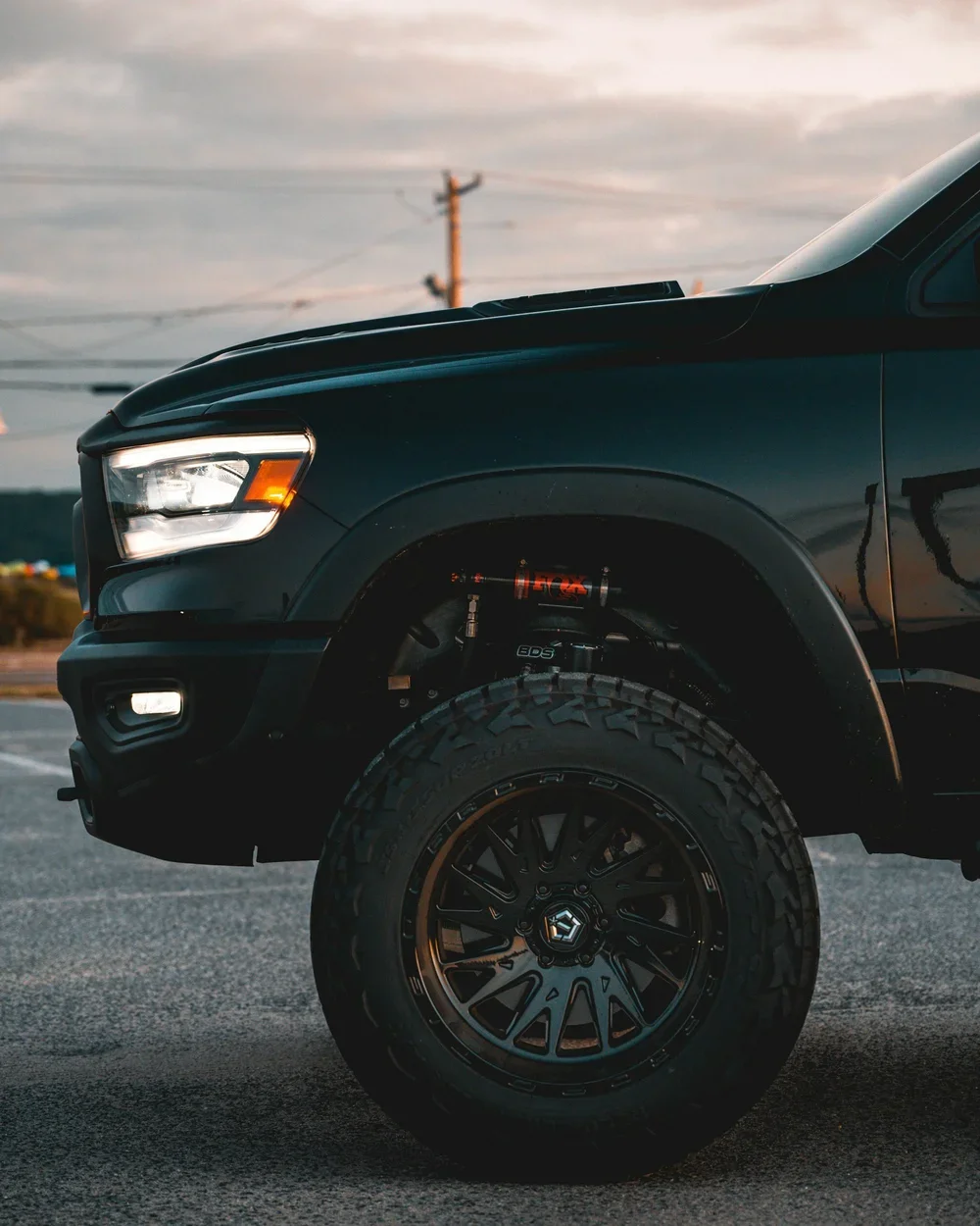Close-up of front part of black pickup truck, showing headlight, tire, and suspension system, parked outdoors during sunset.
