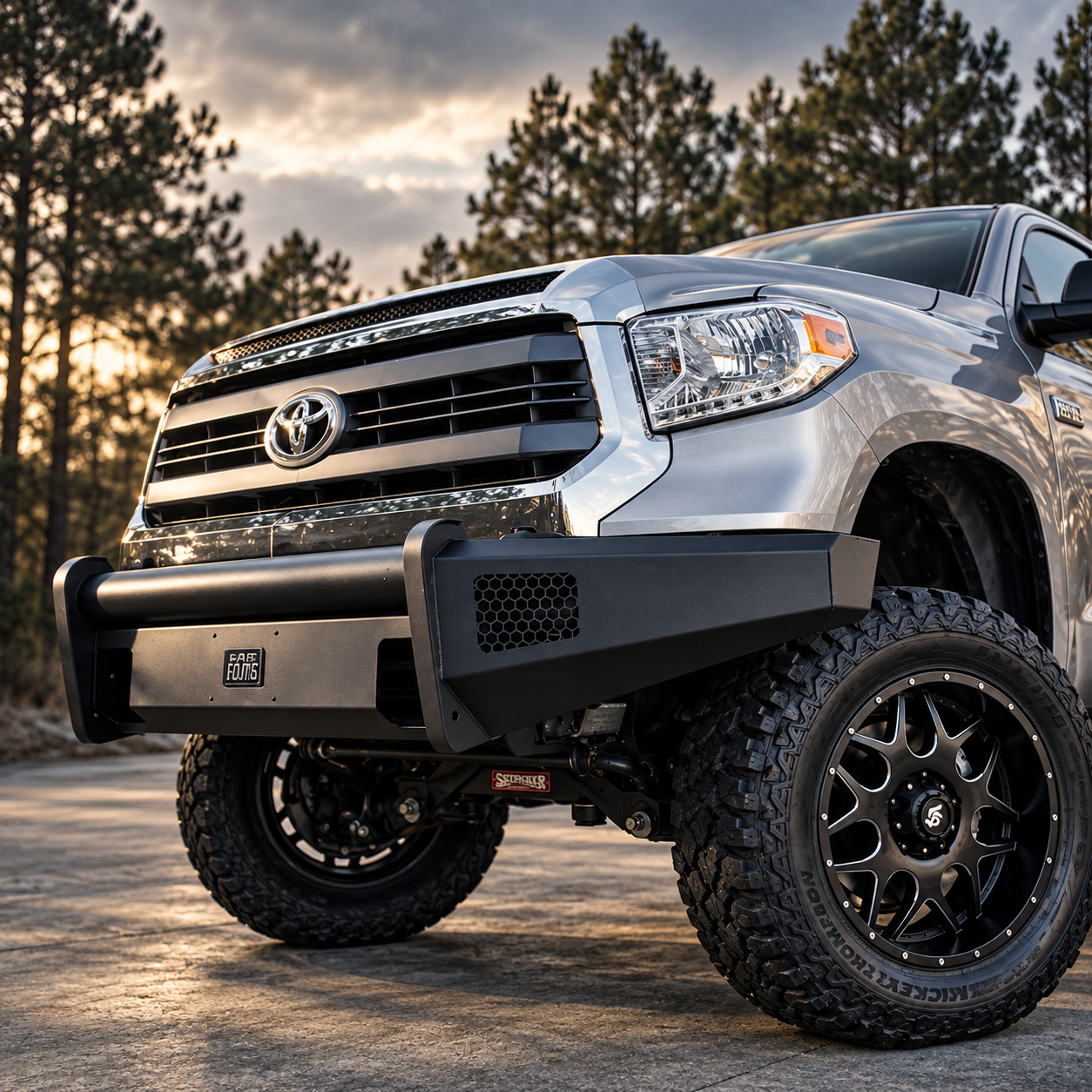 A silver Toyota truck with oversized tires, black off-road bumper, and a protective grille guard parked outdoors during sunset, with pine trees in the background.