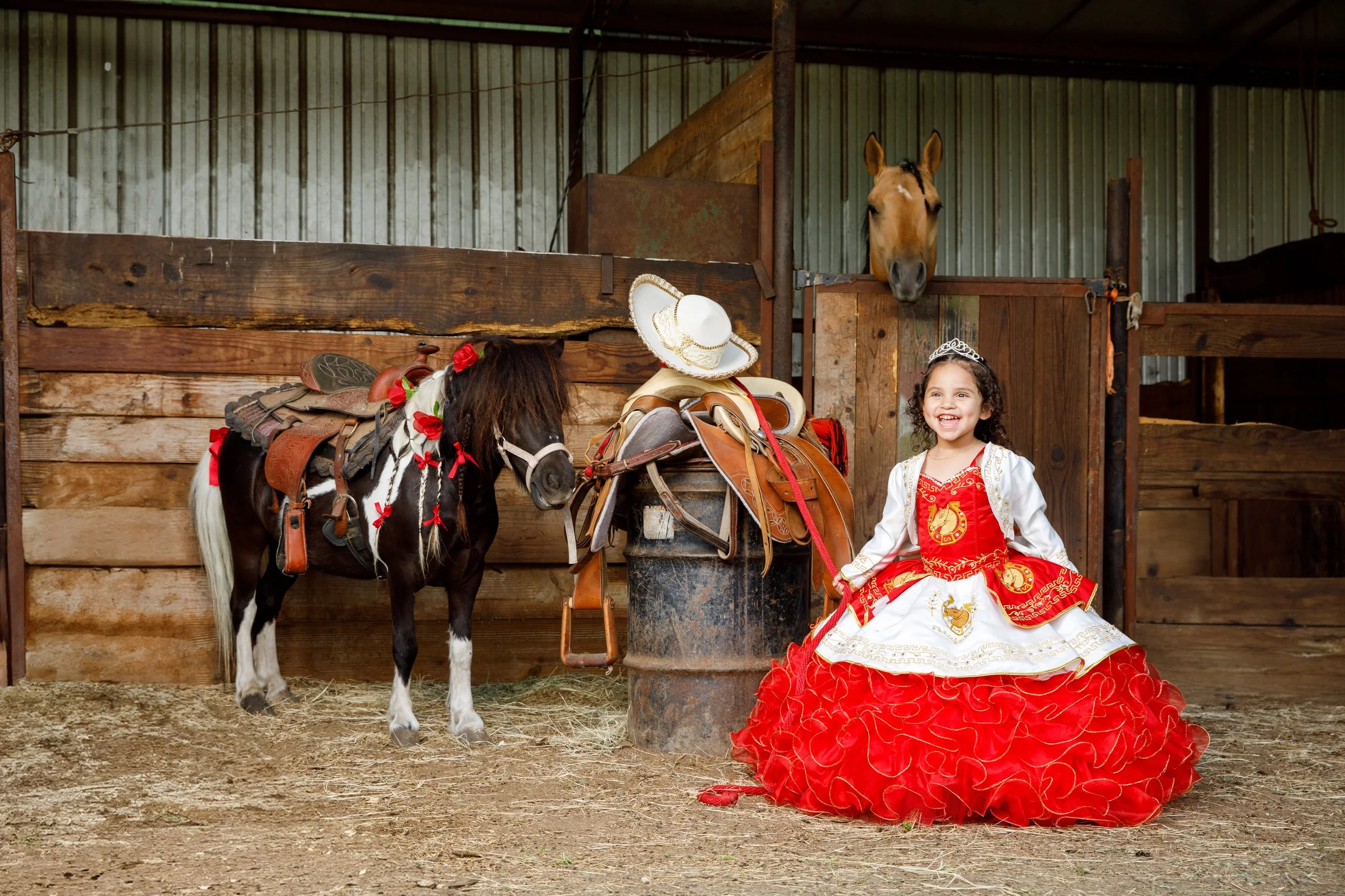 Young girl dressed as a princess sitting on the ground next to a small horse in a stable, with horse tack and a cowboy hat placed on a barrel nearby, and a horse peeking over a stable door.