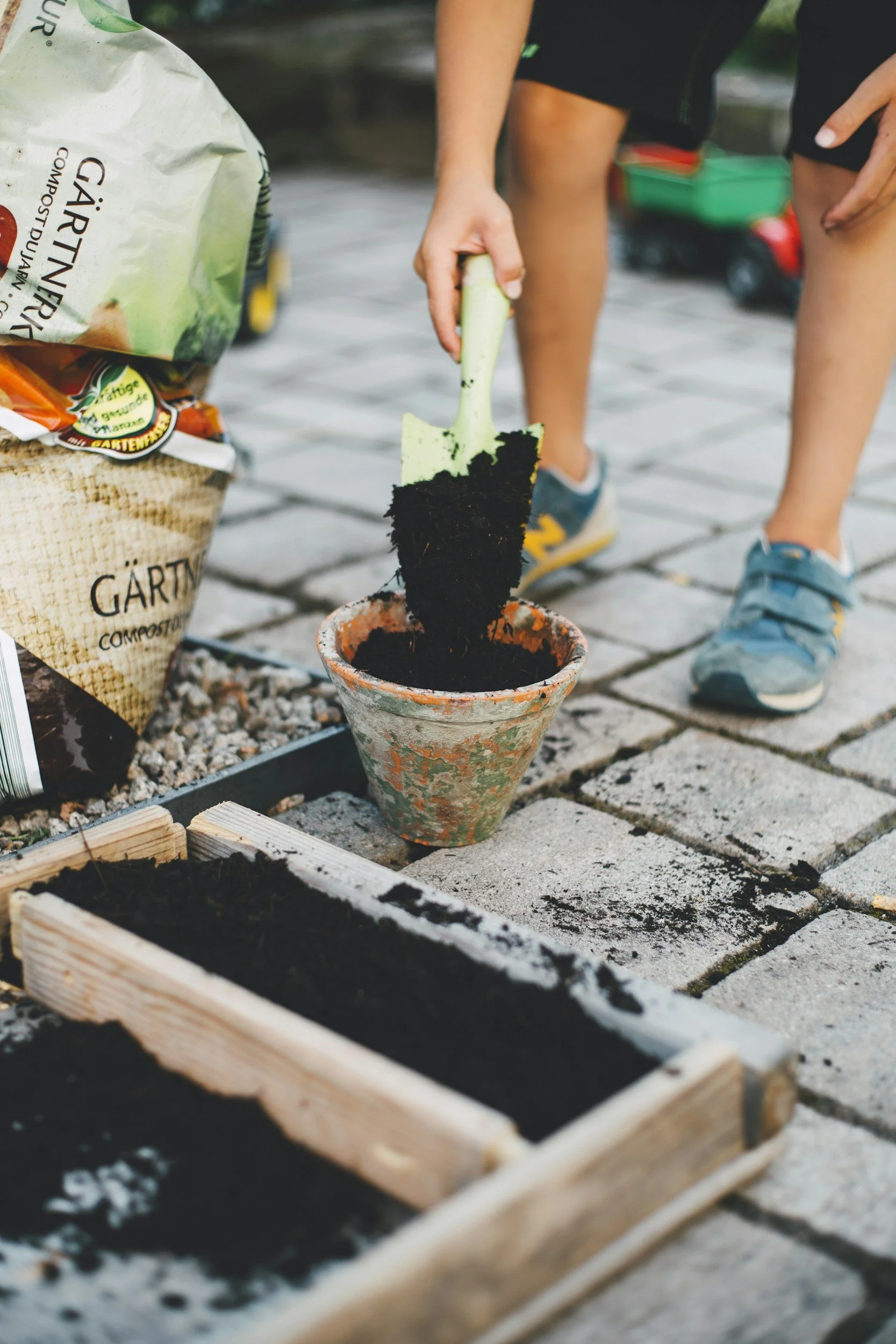 Person planting a seedling in a flowerpot on a stone surface, gardening tools, compost bag, and planting bed visible.