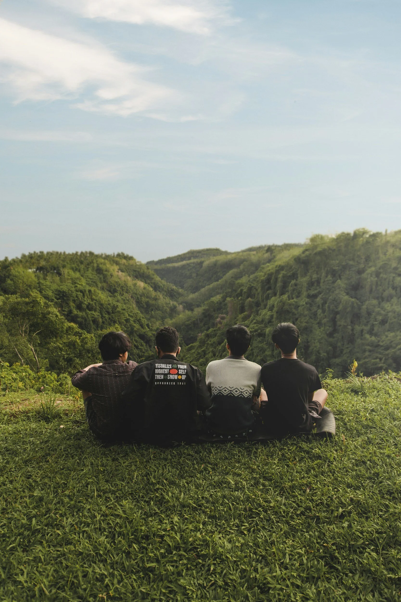 Four young men sit on the grass overlooking a lush green valley with hills covered in dense trees under a partly cloudy sky.