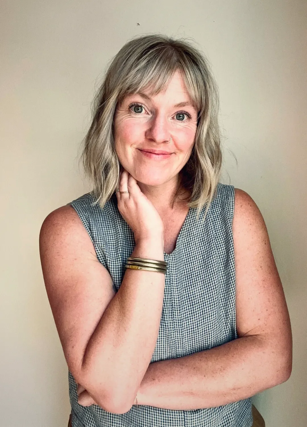 A woman with shoulder-length blonde hair, wearing a sleeveless checkered top, smiling, touching her neck with one hand, standing against a plain beige wall.