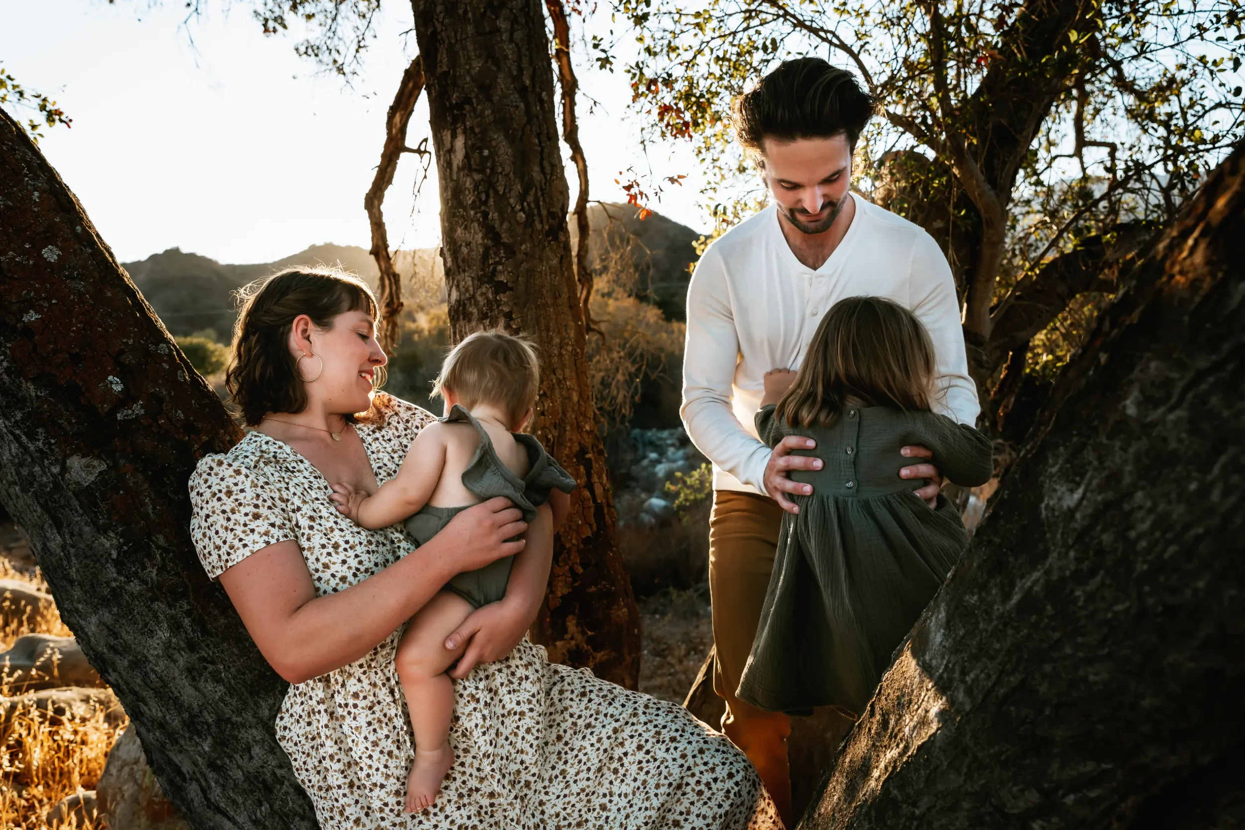 A family of four enjoying time outdoors in a park-like area during sunset. The mother, sitting in a tree, is holding a young child, while another child is hugging a man, possibly the father, who is smiling down at the child. The scene is warm and captures a joyful moment among the family members surrounded by trees.
