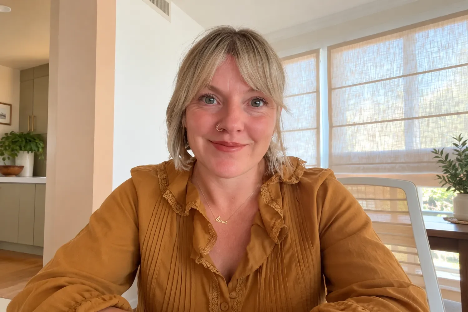 A woman with short blonde hair, wearing a brown top, sitting indoors with a smile, in a room with large windows covered by light blinds, a potted plant, and a kitchen in the background.