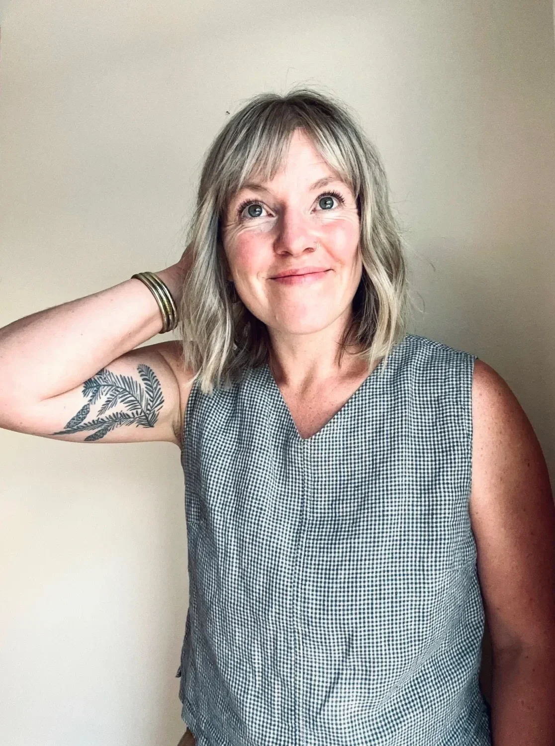 A woman with shoulder-length blonde hair, blue eyes, and a tattoo on her left arm, smiling and looking upward, wearing a sleeveless checkered top and bracelets on her right wrist, standing against a plain beige background.